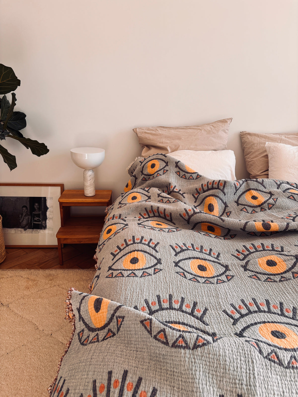 Ein gemütliches Bett mit beigen Kissen und der LEY! Musselin Decke - Vision in Gelb-Blau (2,20 x 2,50 m), neben einem hölzernen Nachttisch mit einer weißen Lampe und einem gerahmten Foto, in einem minimalistischen, Boho-inspirierten Schlafzimmer mit einer Blattpflanze.