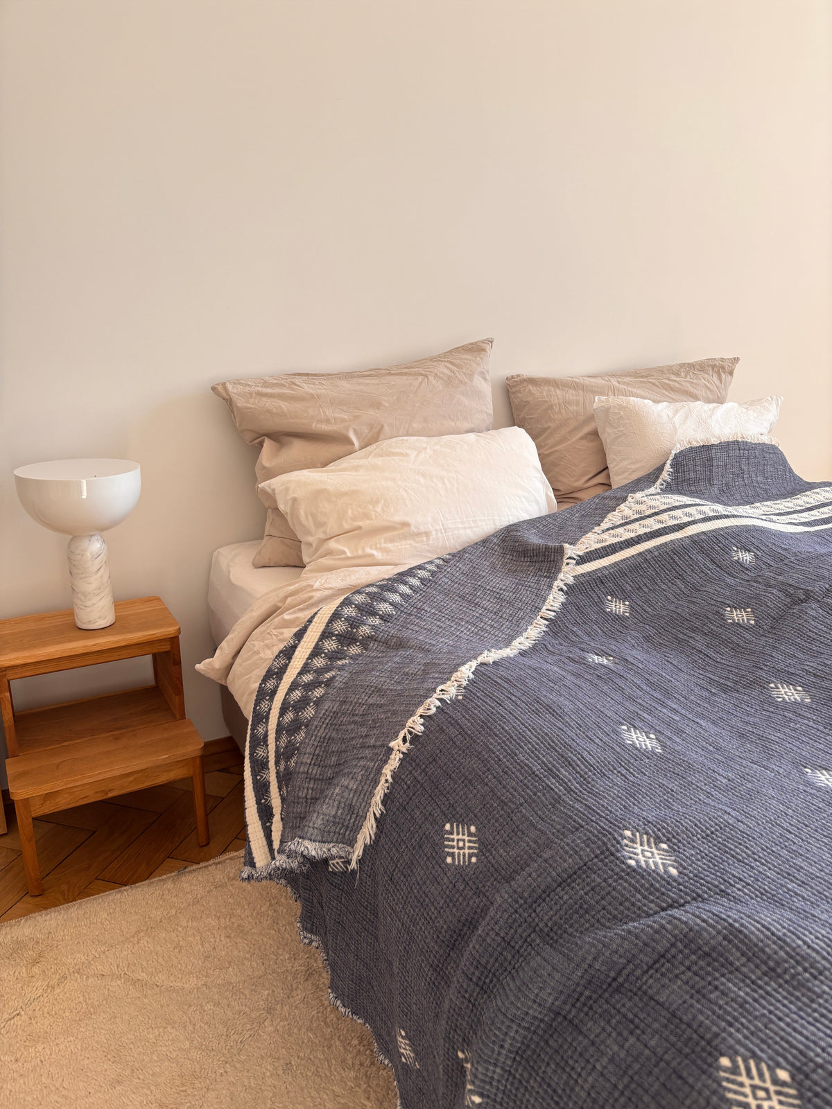Ein ordentlich gemachtes Bett mit beigen Kissen, weißen Laken und einer blau-weißen LEY! Musselin Decke (2,20x2,50m) steht neben einem hölzernen Nachttisch mit einer modernen weißen Lampe in einem gemütlichen, minimalistischen Schlafzimmer.
