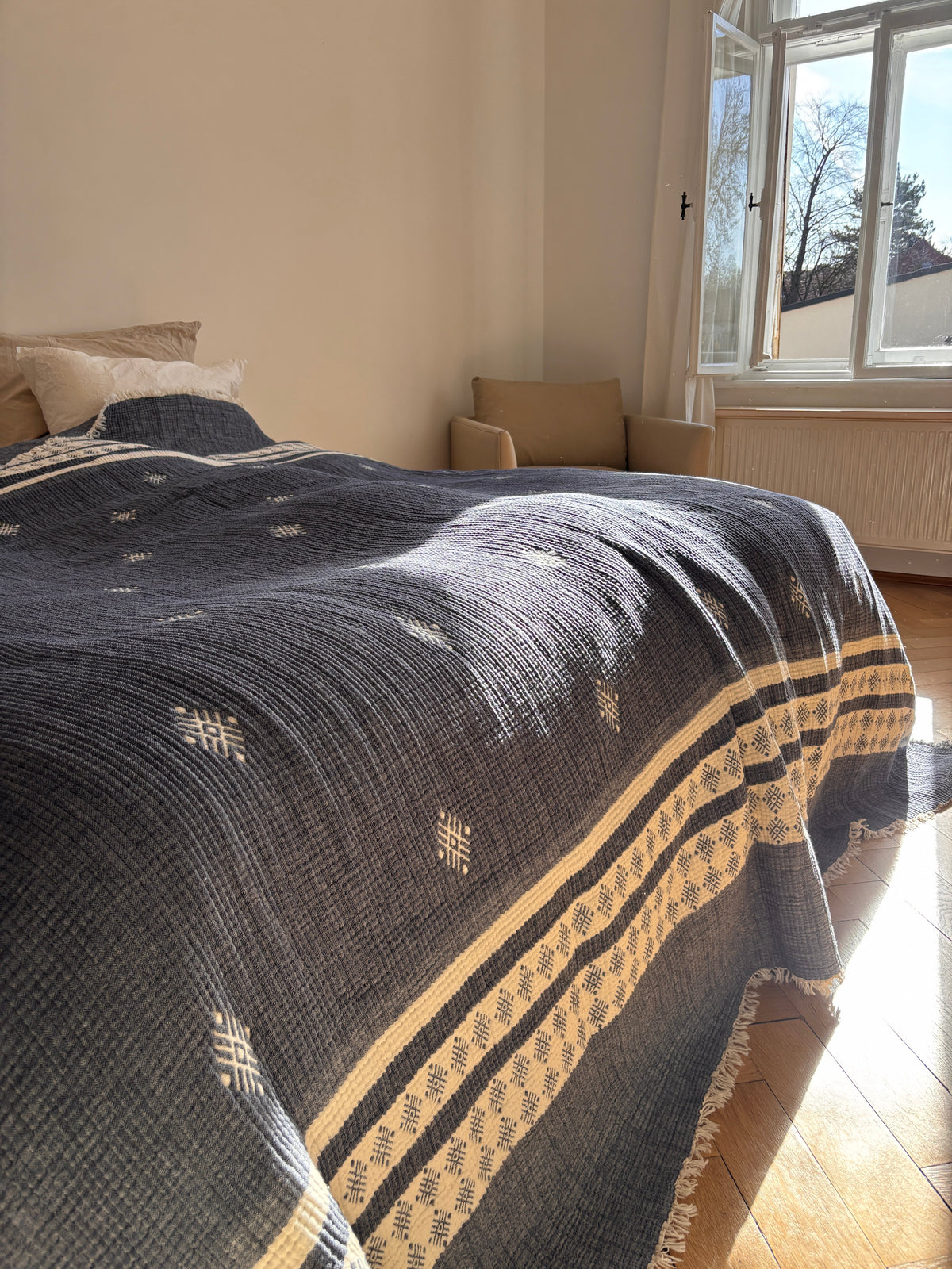 Sonnenlicht erfüllt ein helles, minimalistisches Schlafzimmer, in dem ein ordentlich gemachtes Bett mit der LEY! Musselin Decke - Blau-Weiß (2,20 x 2,50 m). Ein beiger Sessel am Fenster unterstreicht die subtile Boho-Atmosphäre.