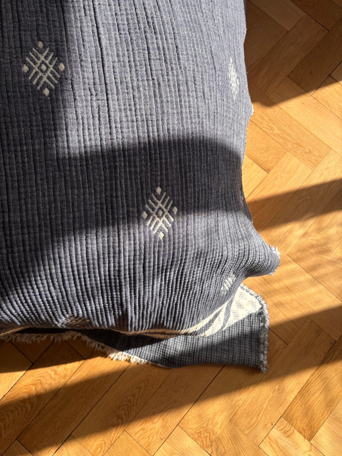 Die LEY! Musselin Decke - Blau-Weiß (2,20 x 2,50 m) liegt teilweise gefaltet auf einem hellen Parkettboden im Sonnenlicht und setzt einen gemütlichen, stilvollen Akzent in jeder Boho-Einrichtung.