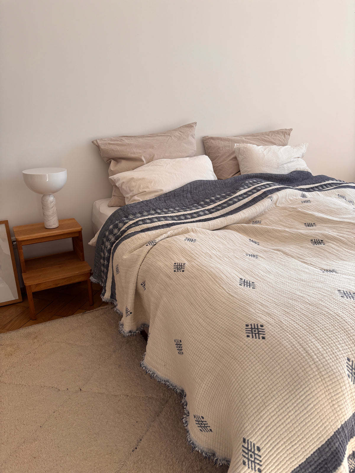 Ein ordentlich gemachtes Bett mit der LEY! Musselin Decke in Blau-Weiß (2,20 x 2,50 m), beiges und blaues Bettzeug und neutrale Kissen. Ein hölzerner Nachttisch mit einer weißen Lampe und einem gerahmten Kunstwerk steht auf einem hellbeigen Teppich neben dem Bett.