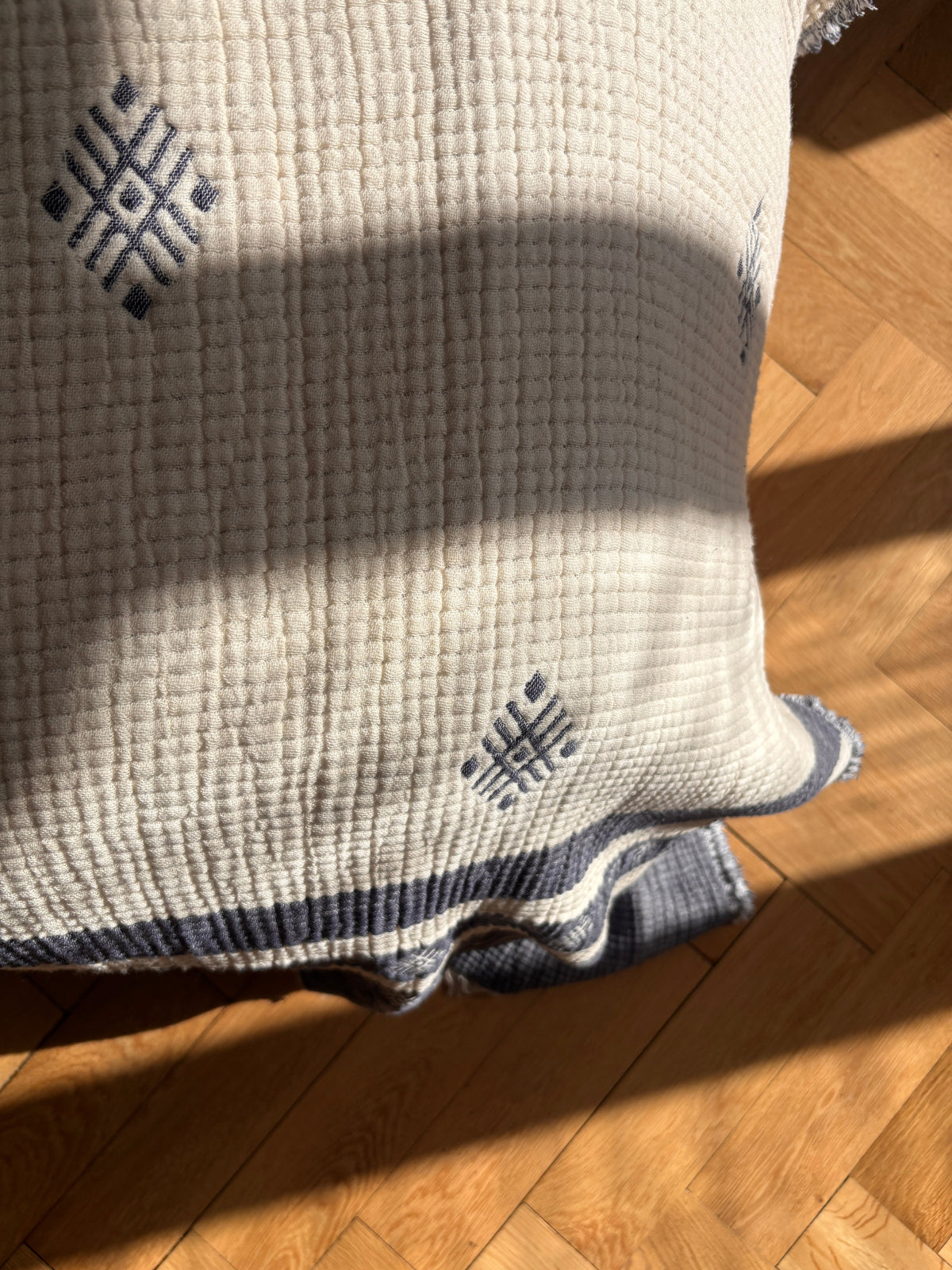 Eine Nahaufnahme der LEY! Musselin Decke - Blau-Weiß (2,20 x 2,50 m) mit blauen geometrischen Mustern und einer gestreiften Kante im Boho-Design, die gemütlich über einen sonnenbeschienenen Holzparkettboden drapiert ist.