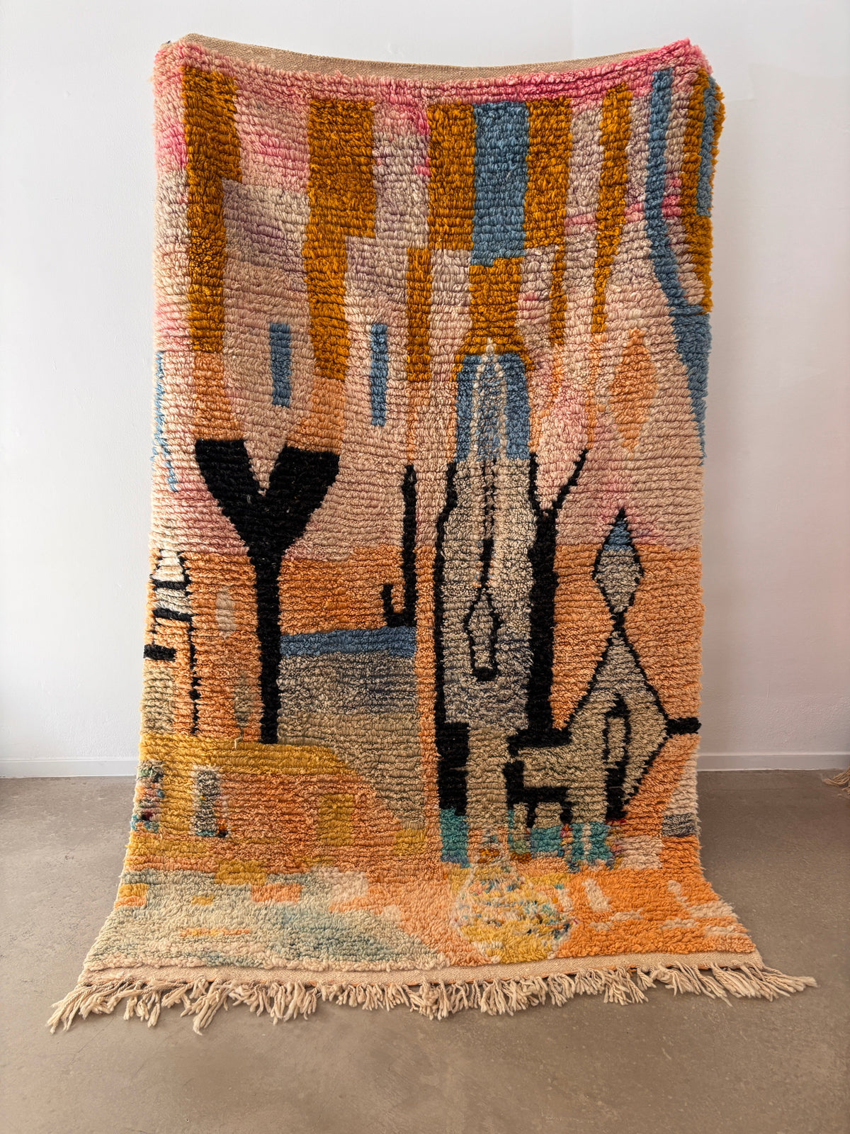 Der LEY! Berber Teppich - Biama (1,60 x 2,60) ist ein farbenfroher, handgewebter Teppich mit abstrakten geometrischen Mustern in rosa, gelber, orangefarbener, blauer und schwarzer Wolle sowie Fransen, der aufrecht vor einer weißen Wand auf einem Betonboden steht.