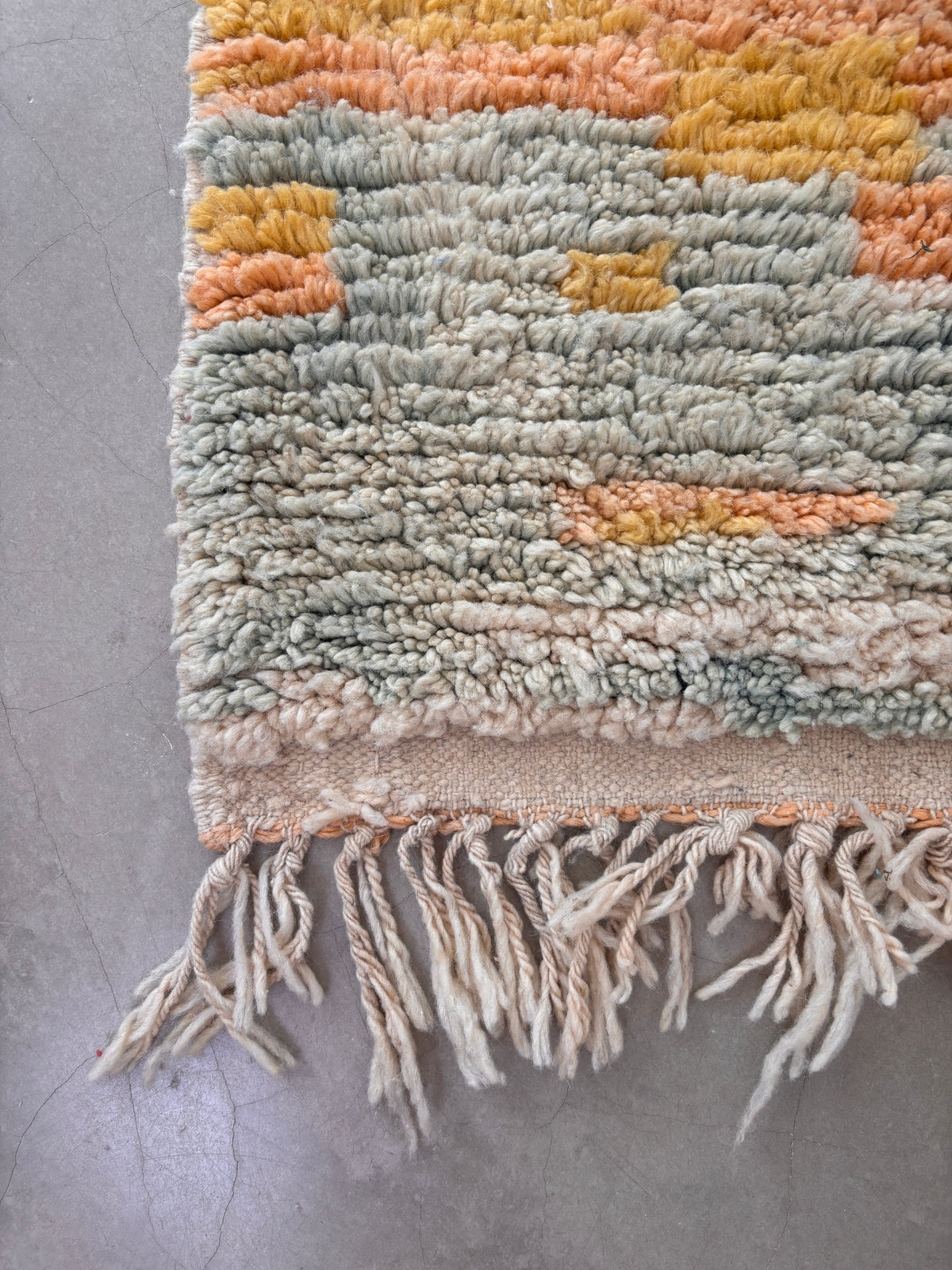 Eine Nahaufnahme der handgefertigten LEY! Berber Teppich - Biama (1,60 x 2,60) aus Schurwolle mit pastellorangen, gelben und blauen Tönen sowie Fransen an der Kante, liegend auf einem hellgrauen Betonboden.