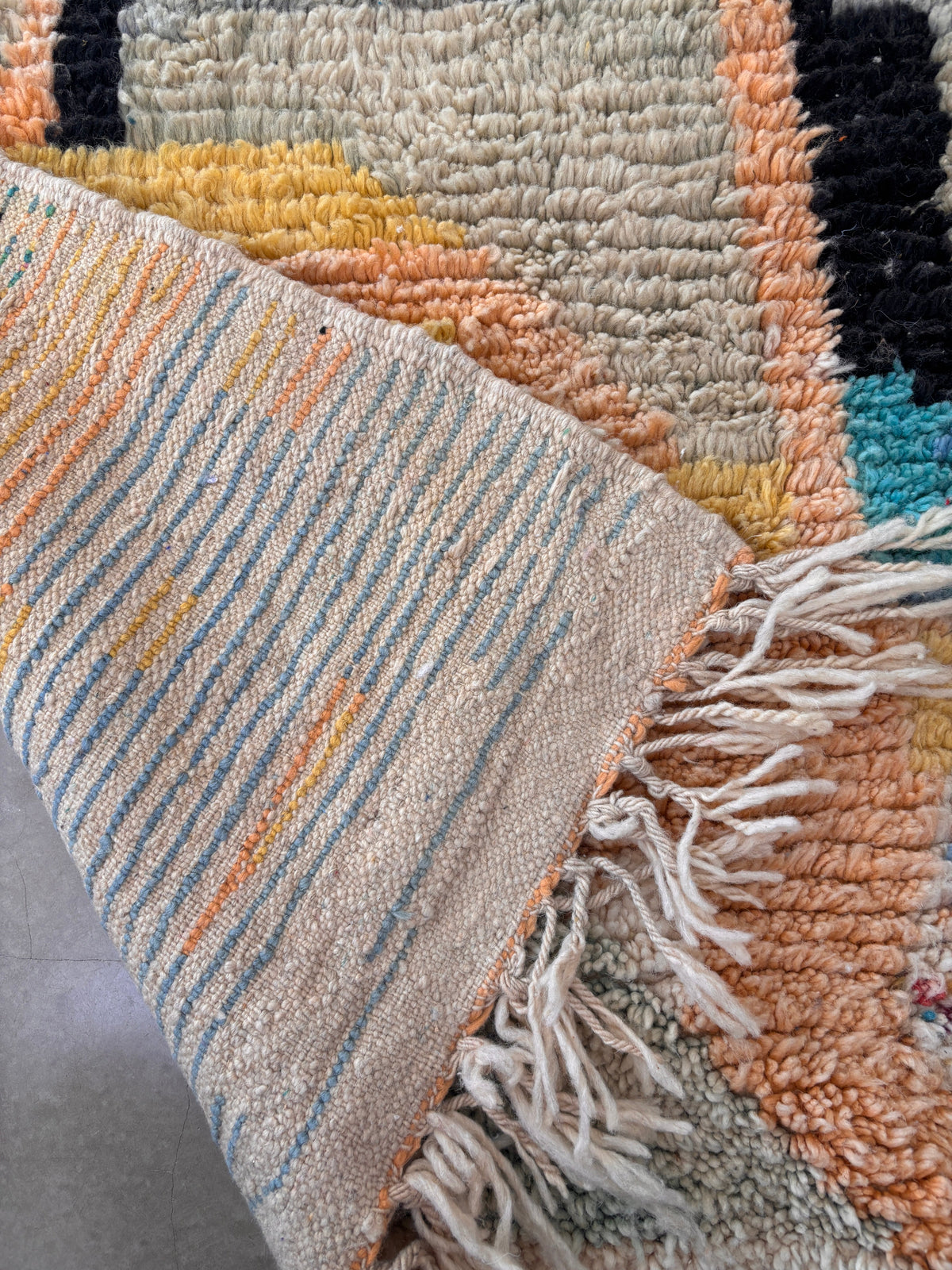 Eine Nahaufnahme des LEY! Berber Teppich - Biama (1,60 x 2,60), handgefertigt mit blauen und gelben Streifen und weißen Fransen auf der Rückseite; pfirsichfarbene, cremefarbene, schwarze und gelbe Schurwolle-Strukturmuster auf der Vorderseite.