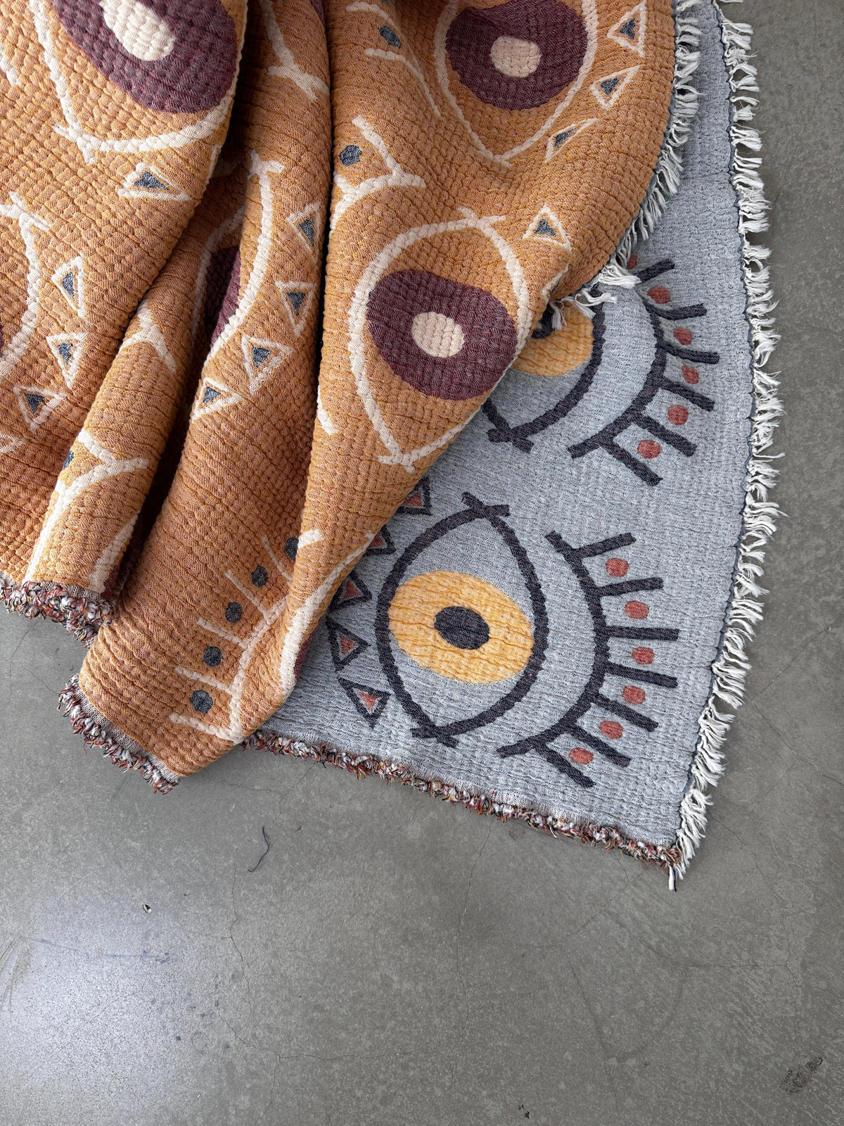 Die LEY! Musselin Decke - Vision - Gelb-Blau (2,20 x 2,50 m) mit kühnem Auge und geometrischen Mustern ist über einen grauen Fransenteppich mit Boho-Design-Motiven drapiert, beides auf einem glatten grauen Boden.