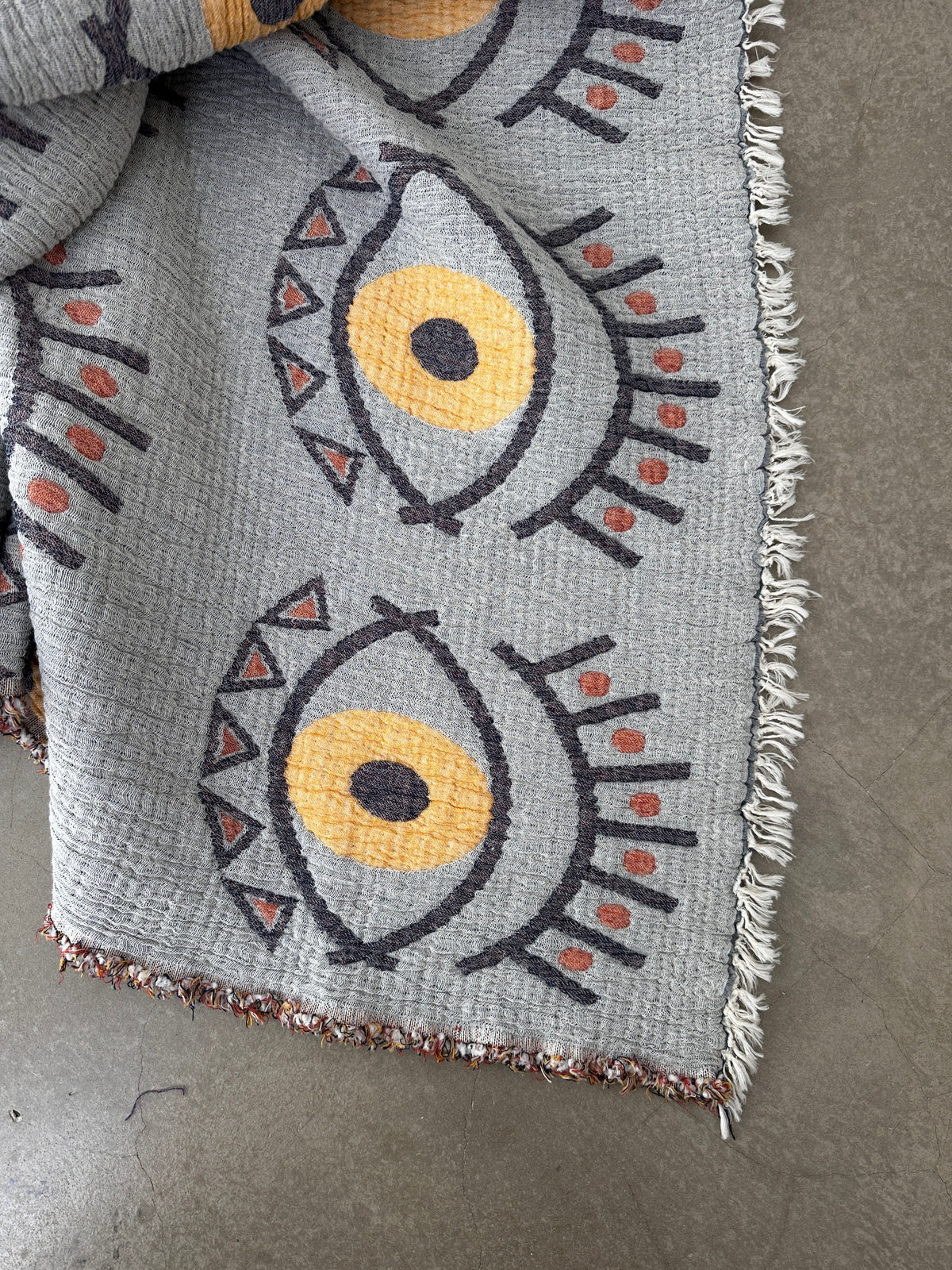 Die LEY! Musselin Decke - Vision - Gelb-Blau (2,20 x 2,50 m) zeigt stilisierte Augen mit gelber Iris und rot-schwarzen Akzenten in einem auffälligen Boho-Design, Fransen an den Rändern und ist auf einem Betonboden drapiert.