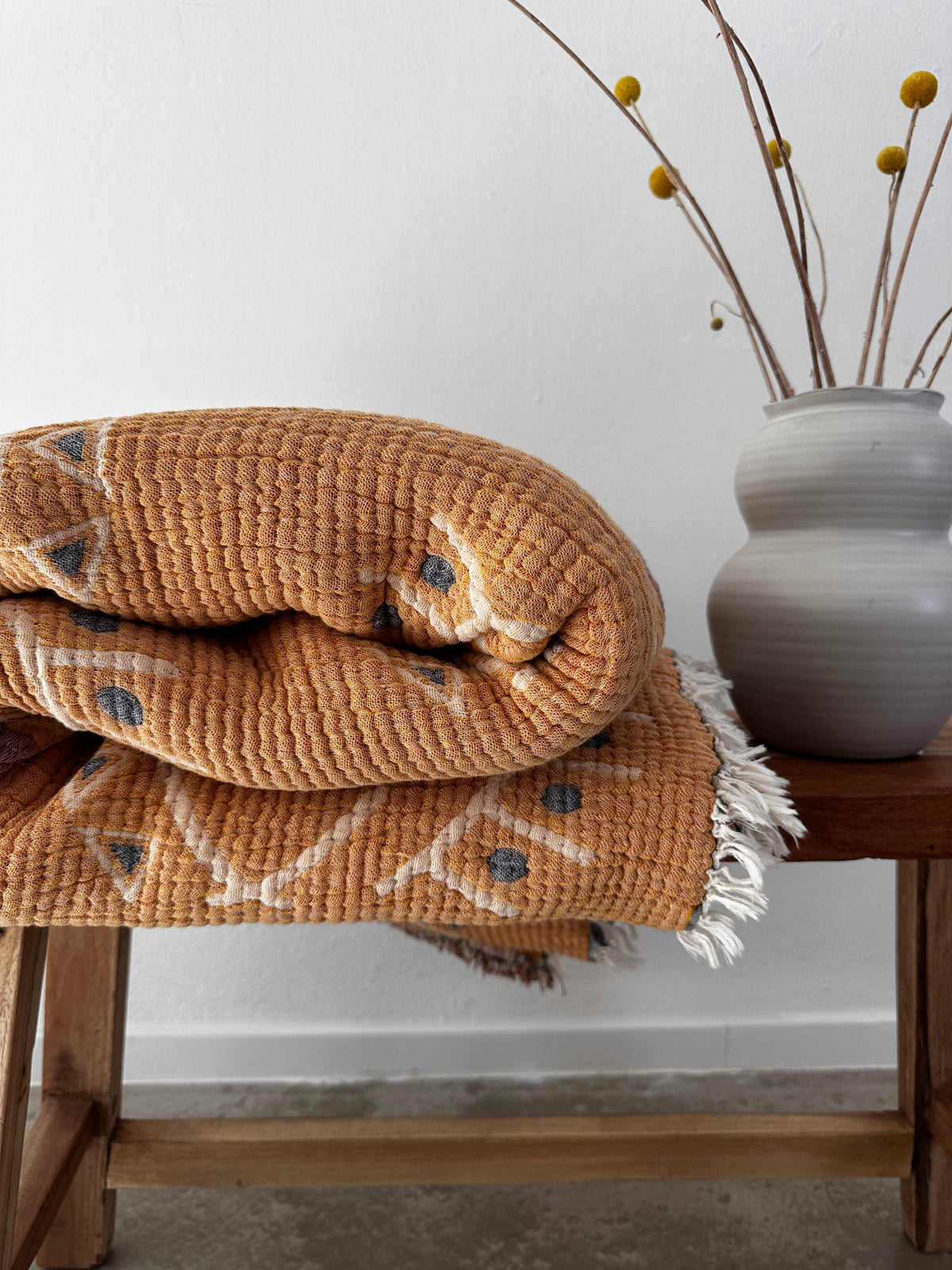 Die LEY! Musselin Decke - Vision - Gelb-Blau (2,20 x 2,50 m), gefaltet mit Fransen, verleiht einer Holzbank neben einer grauen Vase mit gelben Knospenzweigen vor einer weißen Wand einen Boho-Touch.