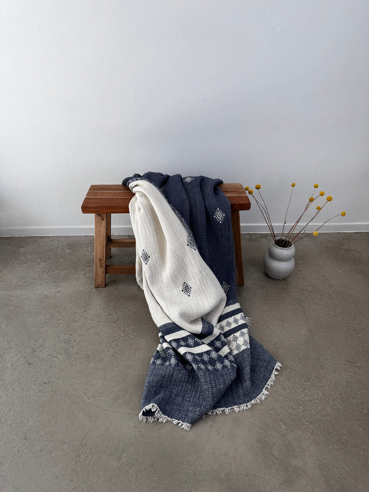 Auf einer Holzbank steht die LEY! Musselin Decke - Blau-Weiß (2,20 x 2,50 m) im Boho-Stil, drapiert neben einer hellgrauen Vase mit gelben Billyball-Blumen auf einem Betonboden vor einer schlichten weißen Wand.