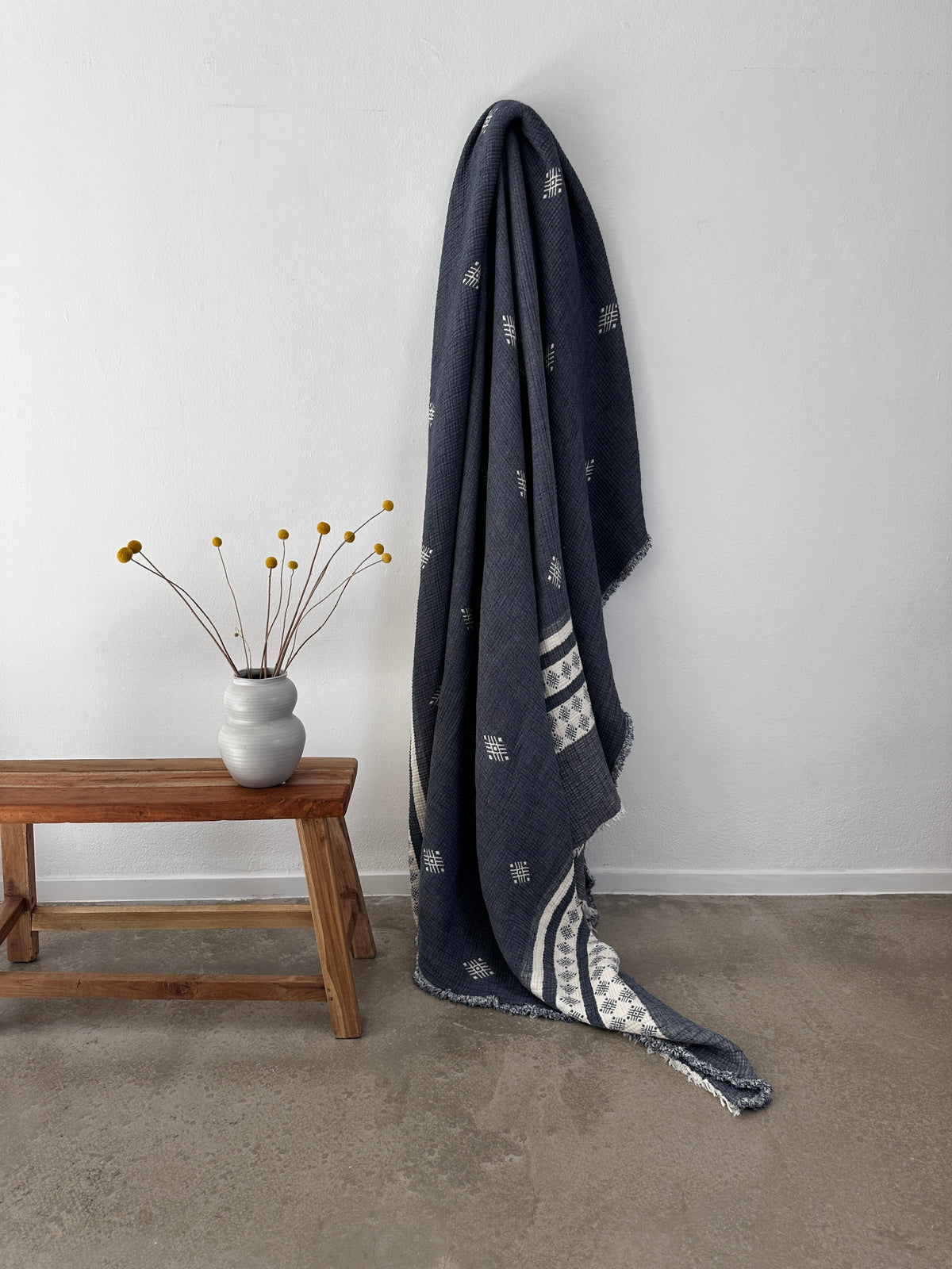 Die LEY! Musselin Decke - Blau-Weiß (2,20 x 2,50 m) im Boho-Design ist an einer weißen Wand neben einer Holzbank drapiert, die eine weiße Vase mit gelben Billy-Ball-Blumen auf einem Betonboden hält.