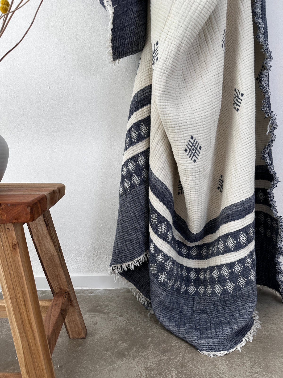 Die Musselin Decke von LEY! (Blau-Weiß, 2,20 x 2,50 m) mit geometrischem Boho-Muster liegt neben einem Holzstuhl auf Betonboden vor einer weißen Wand.
