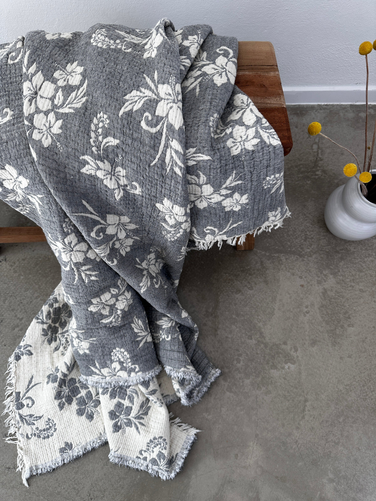 Die Musselin Decke - ROSE von LEY! in Grau-Weiß (2,20 x 2,50 m) mit Boho-Design und Fransen liegt auf einer Holzbank; daneben eine weiße Vase mit gelben Billy Ball Blumen auf poliertem Betonboden.