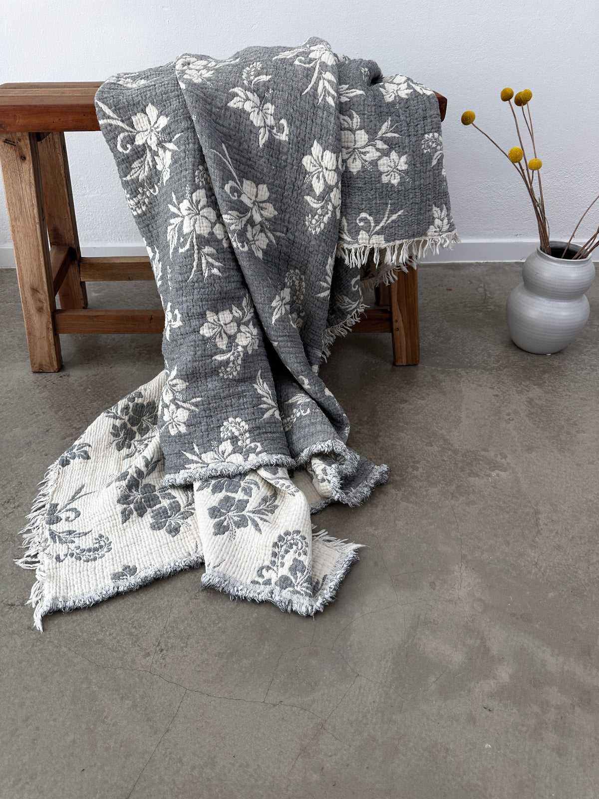 Die Musselin Decke - ROSE von LEY! in Grau-Weiß (2,20 x 2,50 m) mit Boho-Design und Fransen liegt weich über einer Holzbank, ein Ende berührt den Betonboden. Daneben steht eine runde graue Vase mit gelben Billy Balls an der weißen Wand.