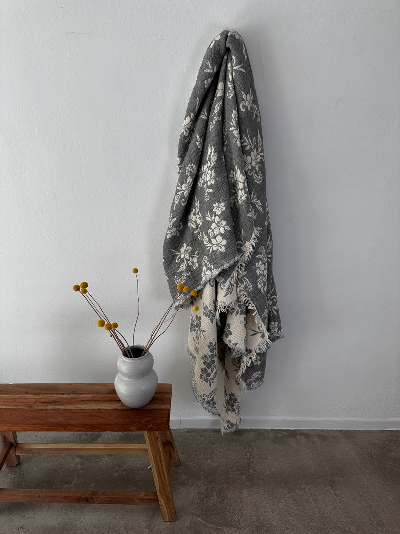 Die Musselin Decke - ROSE von LEY! in Grau-Weiß (2,20 x 2,50 m) mit Boho-Blumenmuster hängt an einer weißen Wand über einer Holzbank. Auf der Bank steht eine Vase mit gelben Billy Balls, der Boden ist grauer Beton.