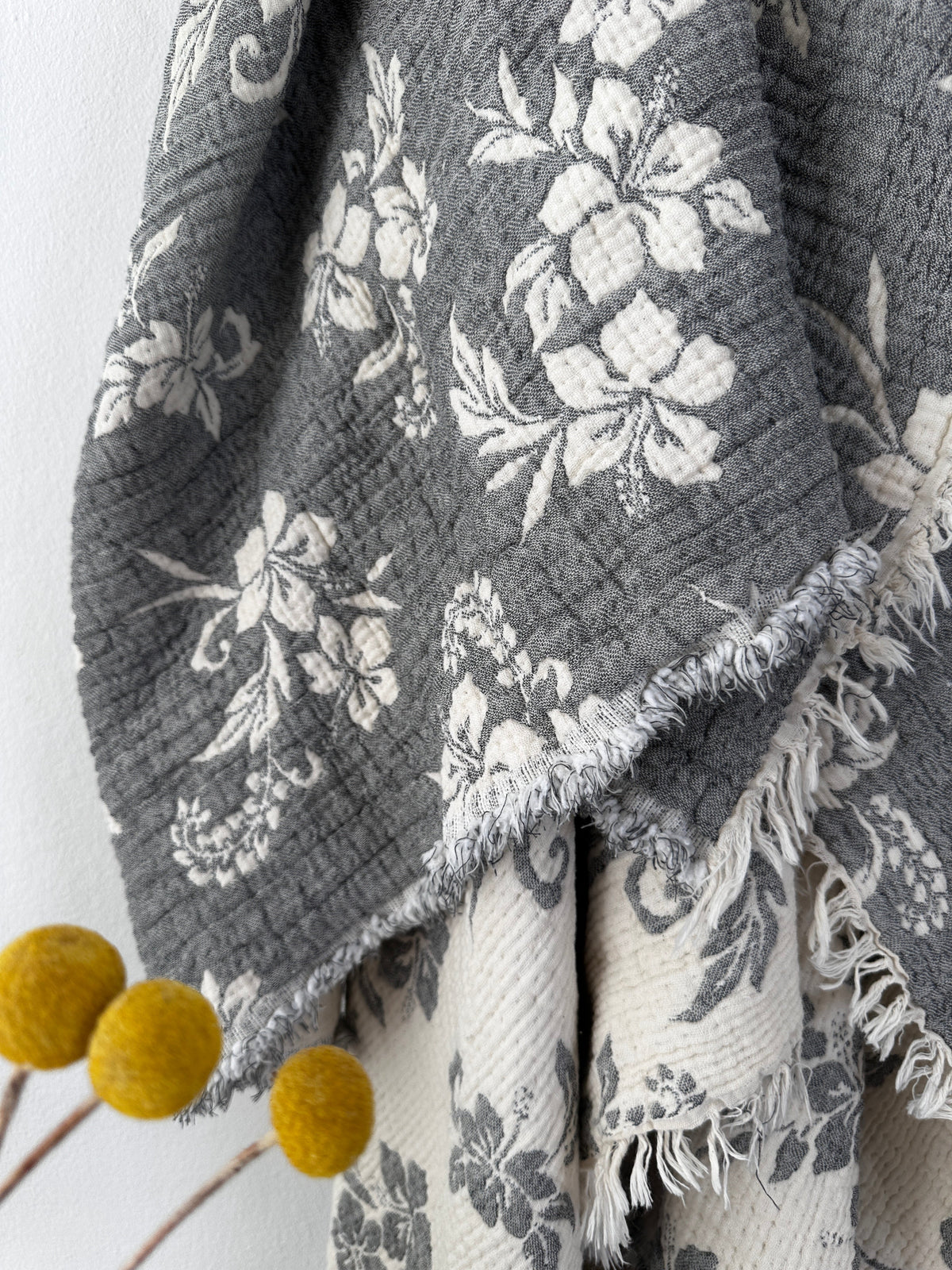 Eine Nahaufnahme von zwei drapierten Stoffen, darunter die LEY! Musselin Decke - ROSE in Grau-Weiß (2,20 x 2,50 m) mit weißen Blumenmustern und Fransen. Der cremefarbene Stoff mit grauen Mustern und gelben Billy-Ball-Blumen verleiht ihm einen Boho-Touch.