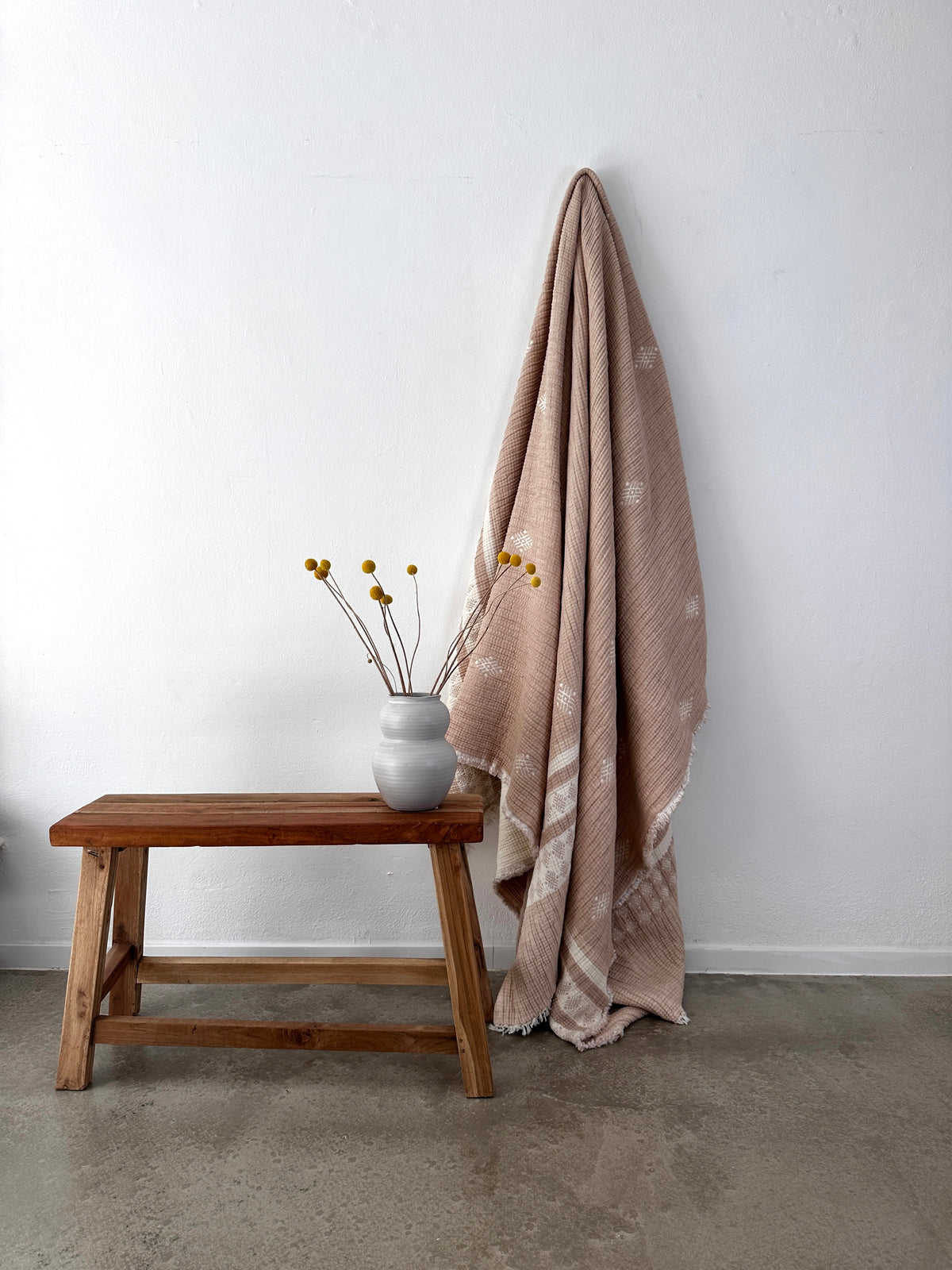 Auf einer Holzbank steht eine weiße Vase mit gelben Billy-Ball-Blumen, daneben hängt die LEY! Musselin Decke - Beige-Weiß (2,20 x 2,50 m) im Boho-Design, die an einer weißen Wand über einem Betonboden hängt.