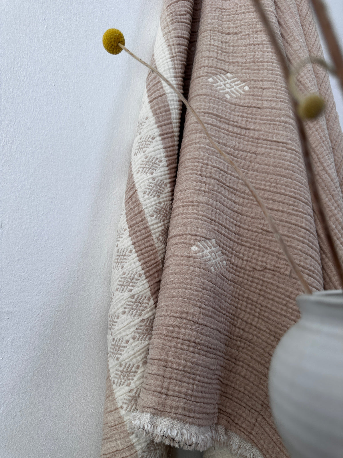 Die Musselin Decke in Beige-Weiß (2,20 x 2,50 m) von LEY! zeigt weiße geometrische Muster - ideal für Boho-Design-Fans. Sie wird hängend neben einer weißen Vase mit dünnen Stielen und kleinen gelben Blumen präsentiert.