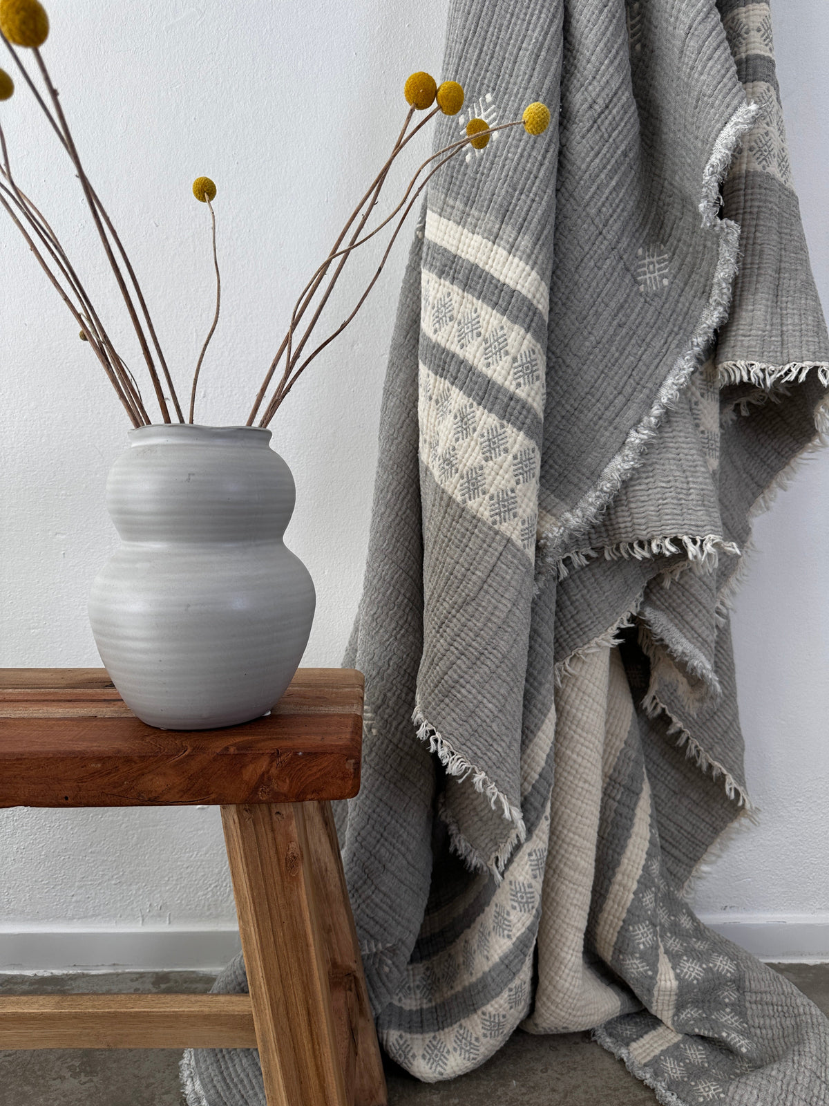 Eine graue Keramikvase mit gelben Billyball-Blüten steht auf einer Holzbank neben der LEY! Musselin Decke - Hellgrau-Weiß (2,20 x 2,50 m), deren ausgefranste Ränder im Boho-Stil vor einer weißen Wand drapiert sind.