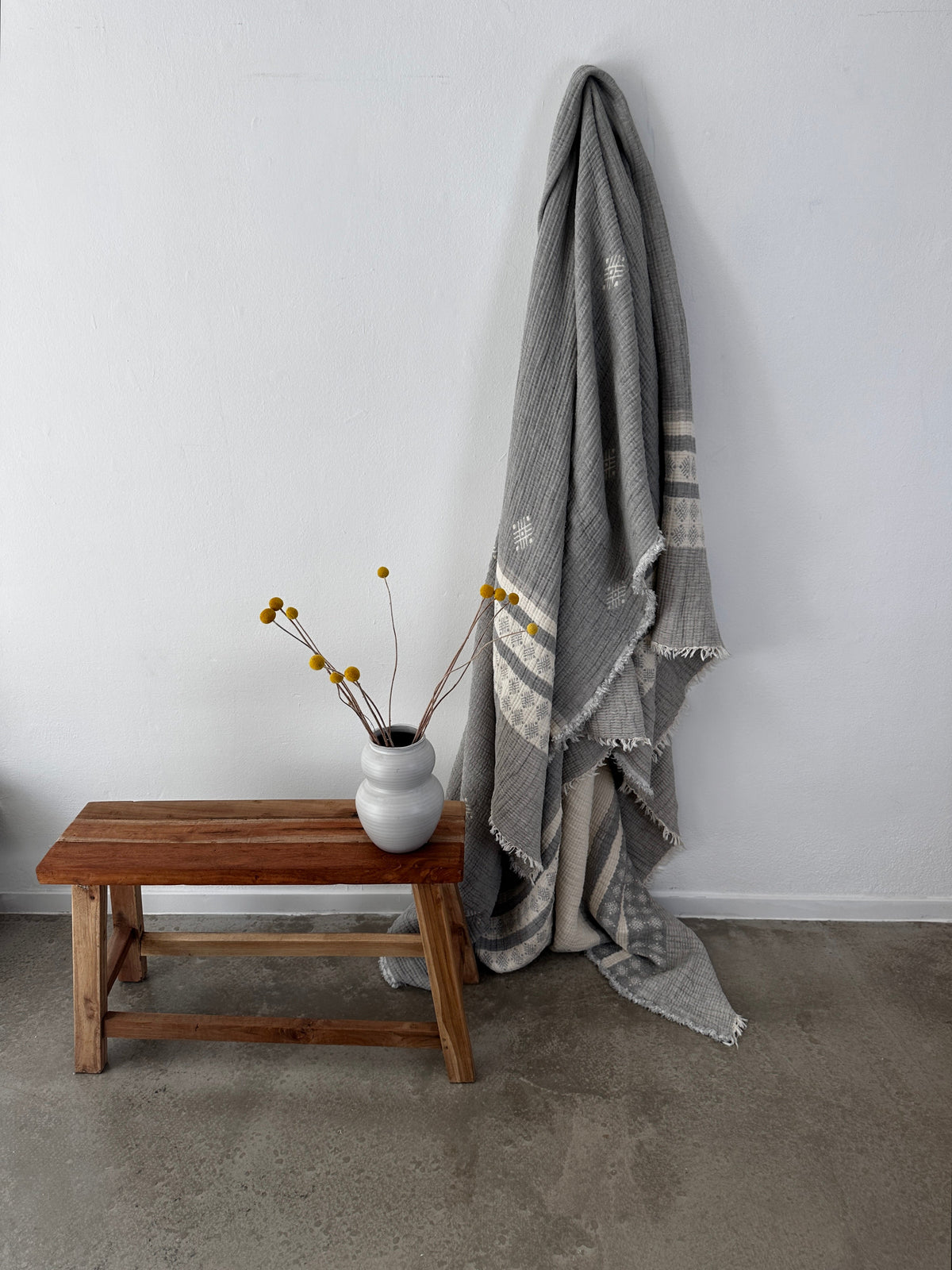 Eine Musselin Decke in Hellgrau-Weiß von LEY! (2,20 x 2,50 m) im Boho-Stil liegt vor einer weißen Wand. Daneben steht eine kleine Holzbank mit einer weißen Vase und gelben Billy Ball Blumen auf glattem Betonboden.