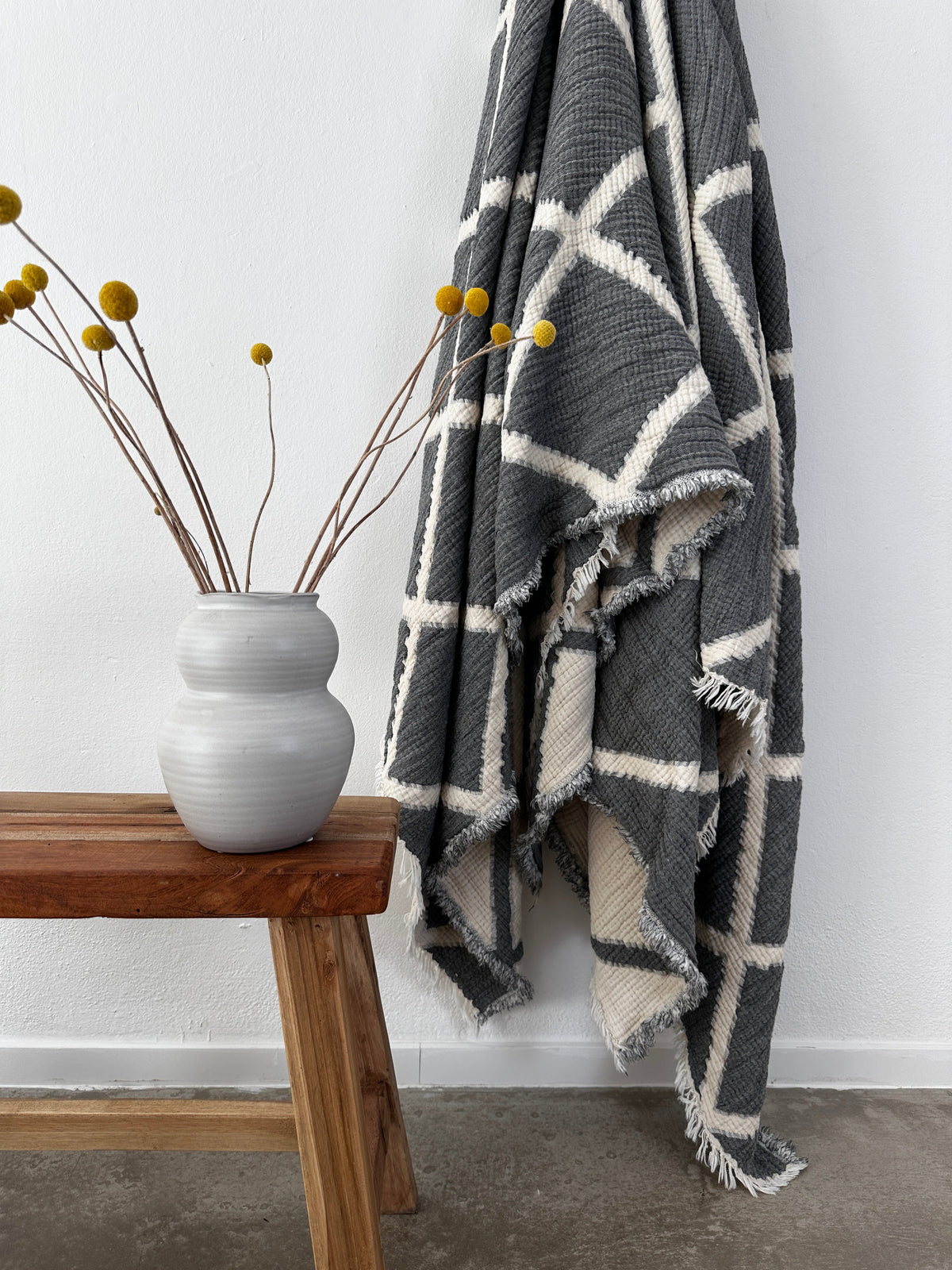 Die Musselin Decke - Grau-Weiß, 2,20 x 2,50 m von LEY! hängt an einer weißen Wand neben einer Holzbank mit gelben Blumen und schafft eine moderne, minimalistische und atmungsaktive Atmosphäre.