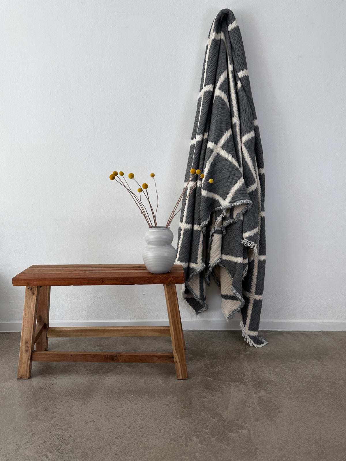 Eine Holzbank mit einer weißen Vase mit gelben Billybällen steht neben der LEY! Musselin Decke - Grau-Weiß (2,20 x 2,50 m) im Boho-Design, drapiert an einer weißen Wand über einem polierten Betonboden.