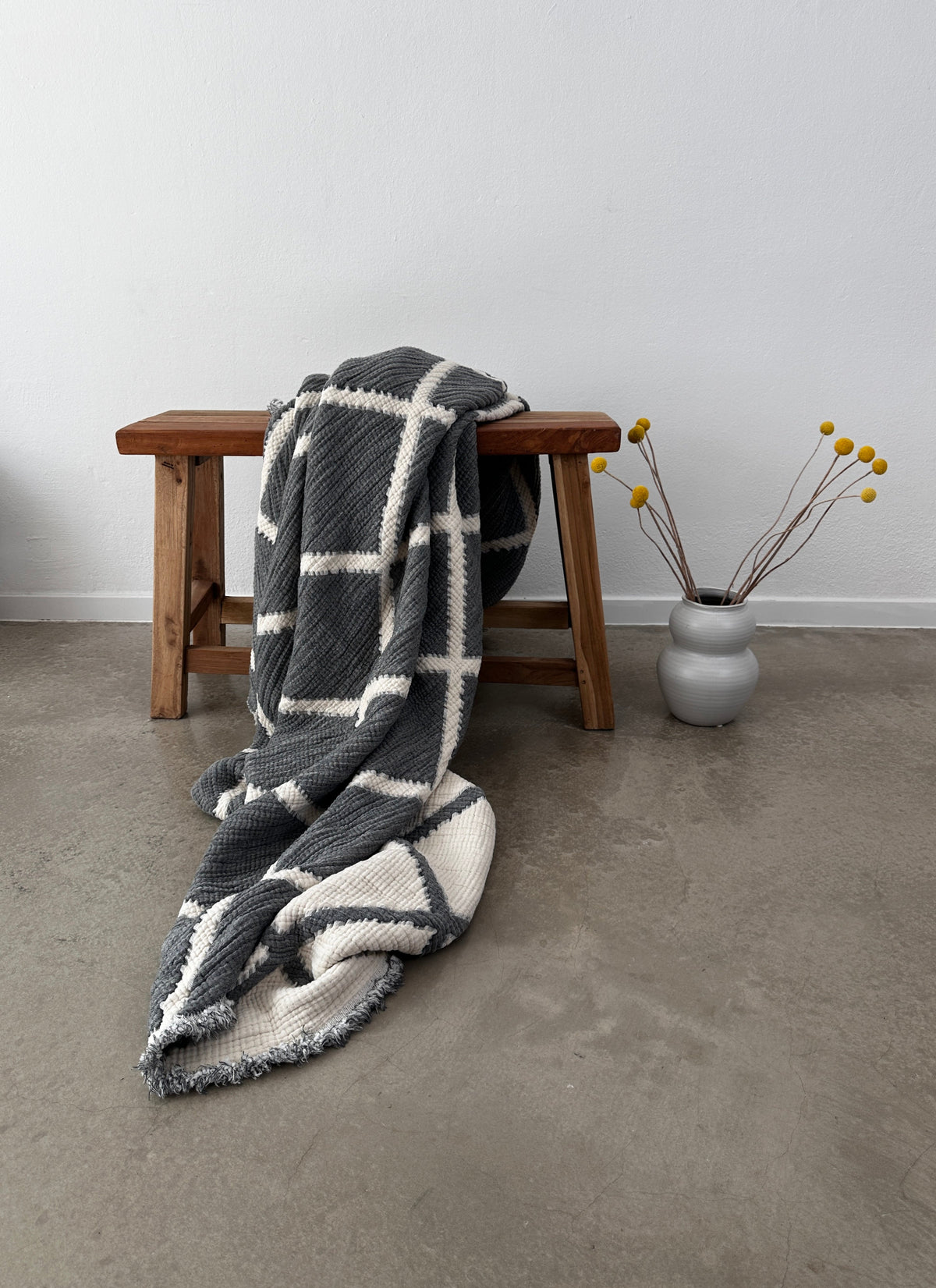 Die Musselin Decke - Grau-Weiß - 2,20 x 2,50 m von LEY! im Boho-Design liegt auf einer Holzbank auf Betonboden, daneben eine graue Vase mit gelben Billy Ball-Blumen vor weißer Wand.