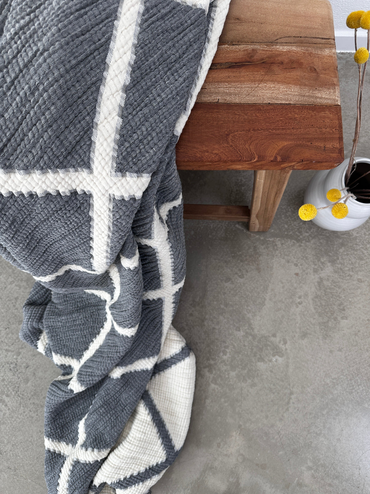 Die LEY! Musselin Decke in Grau-Weiß (2,20 x 2,50 m), atmungsaktiv und im Boho-Design, liegt auf einem Holzstuhl auf Betonboden neben einer weißen Vase mit gelben Billy Balls.