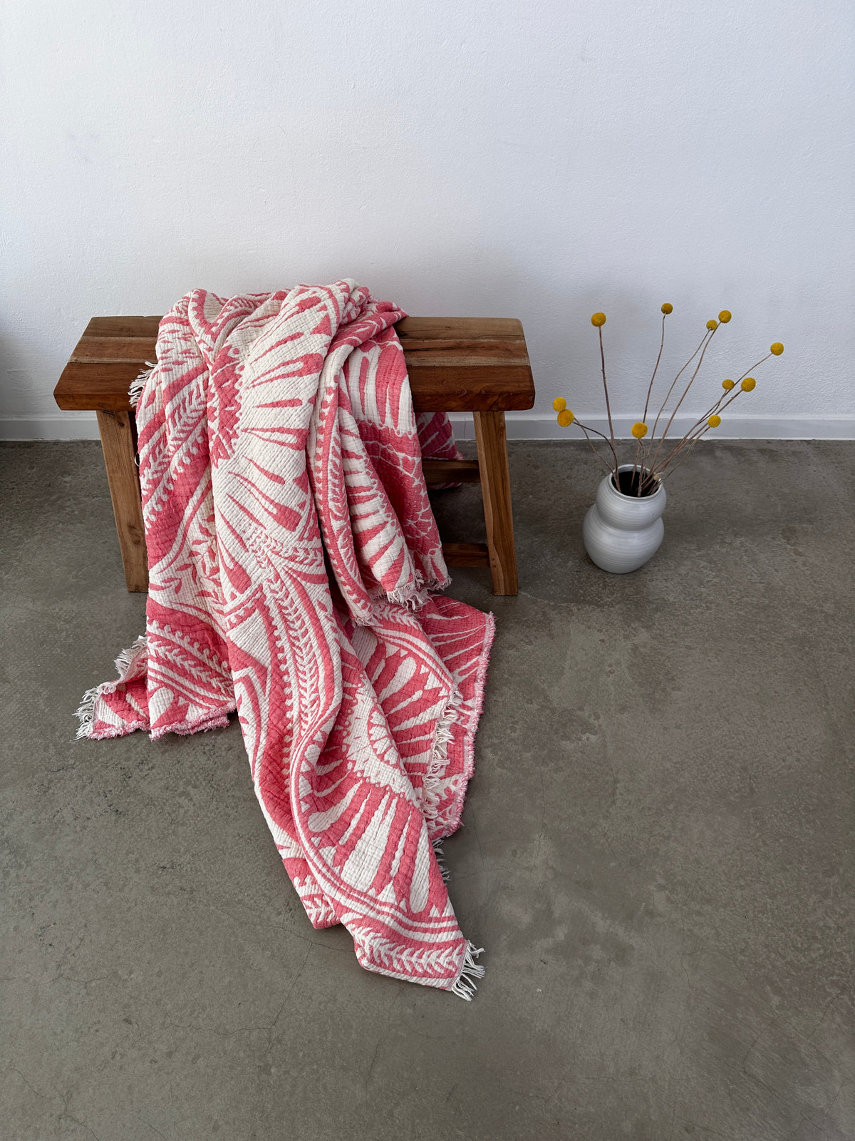 Auf einer Holzbank liegt eine gefaltete CALMA Musselin Decke - Koralle (2,20 x 2,30 m) von LEY! im Boho-Design, Teil der luftigen Korallendecke, die auf dem Boden drapiert ist. Eine weiße Vase mit gelben Bällen steht in der Nähe vor einer schlichten weißen Wand.