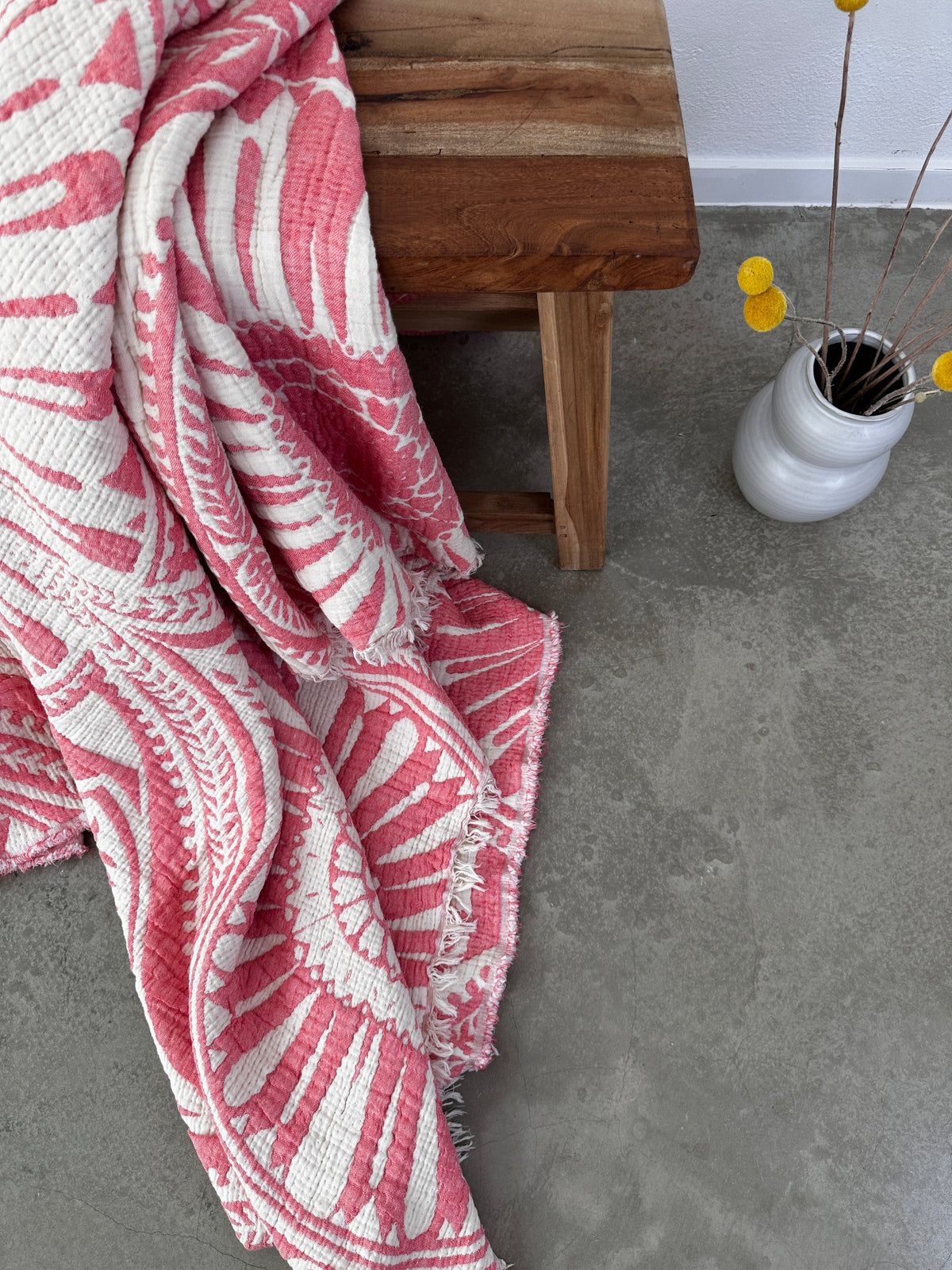 Die CALMA Musselin Decke in Koralle (2,20 x 2,30 m) von LEY! im Boho-Design ist über eine Holzbank auf grauem Betonboden drapiert, daneben steht eine weiße Vase mit gelben Ballblüten.