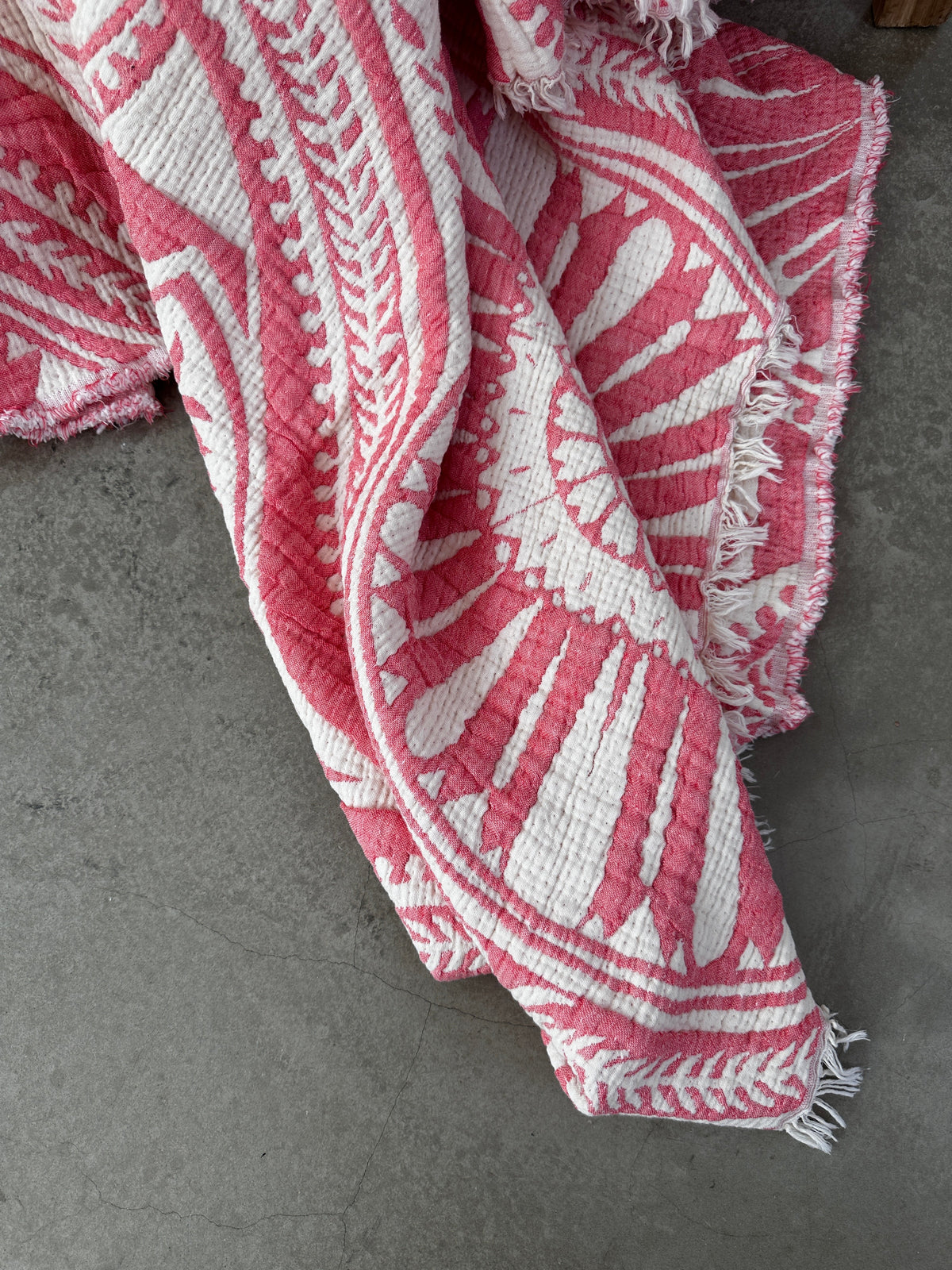 Die CALMA Musselin Decke - Koralle von LEY! (2,20 x 2,30 m) besticht durch ein strukturiertes rosa-weißes Boho-Design mit auffälligen geometrischen und floralen Mustern. Gefranste Kanten unterstreichen die atmungsaktive Decke, die elegant auf einem grauen Betonboden präsentiert wird.
