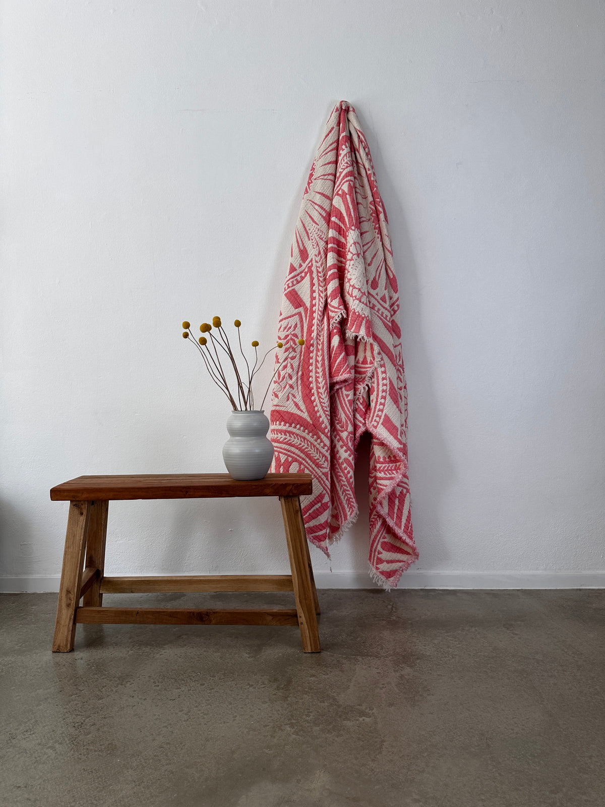 Eine Holzbank mit einer runden weißen Vase mit gelben Billy-Ball-Blumen steht an einer weißen Wand. Darüber hängt die LEY! CALMA Musselin Decke in Koralle (2,20 x 2,30 m), die einen luftigen, atmungsaktiven Boho-Touch verleiht. Der Boden ist aus poliertem Beton.