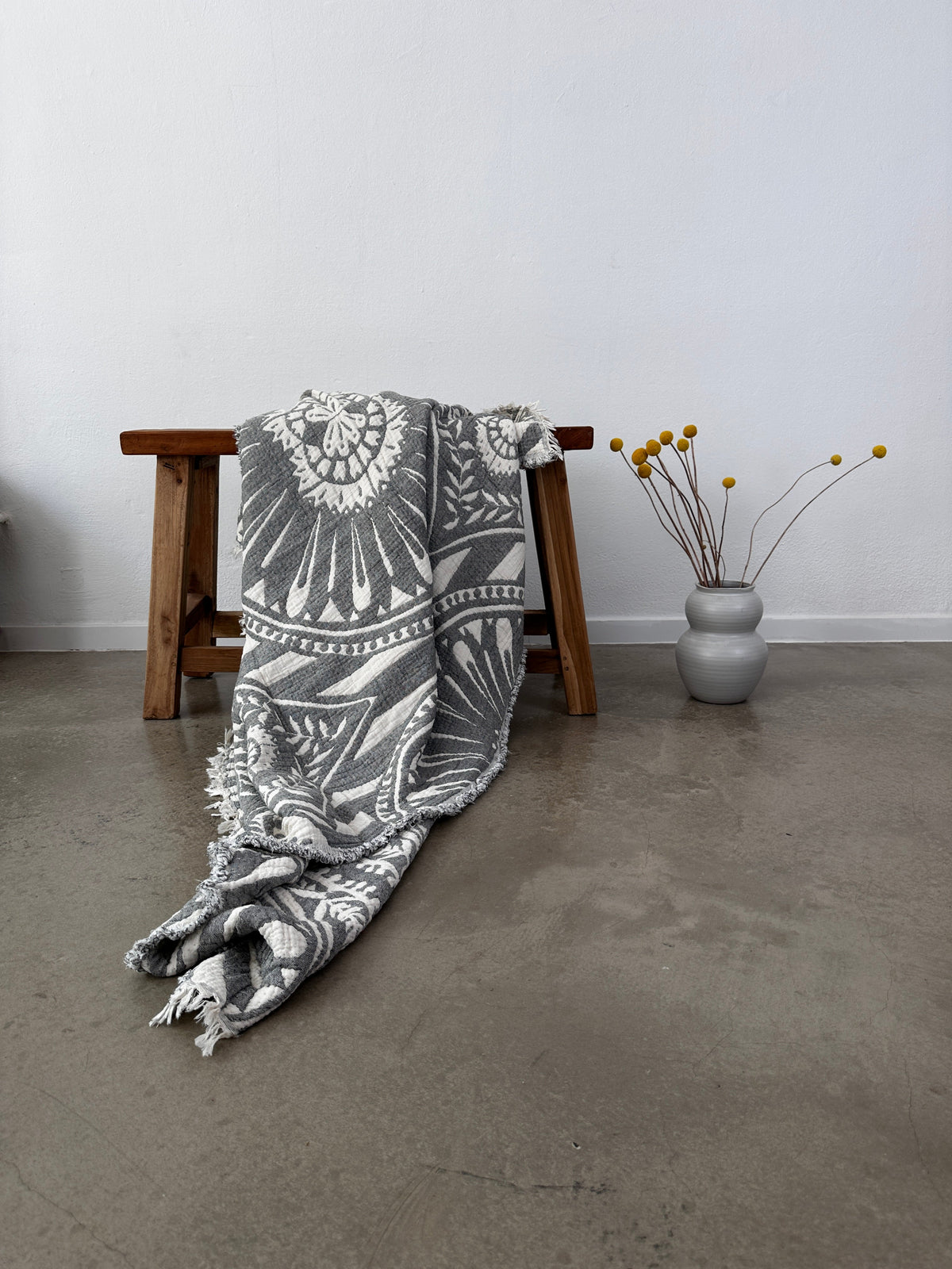 Die CALMA Musselin Decke von LEY! in Dunkelgrau (2,20 x 2,30 m) liegt im Boho-Stil über einer Holzbank auf Betonboden. Daneben steht eine graue Vase mit gelben Billy Ball Blumen vor einer weißen Wand.
