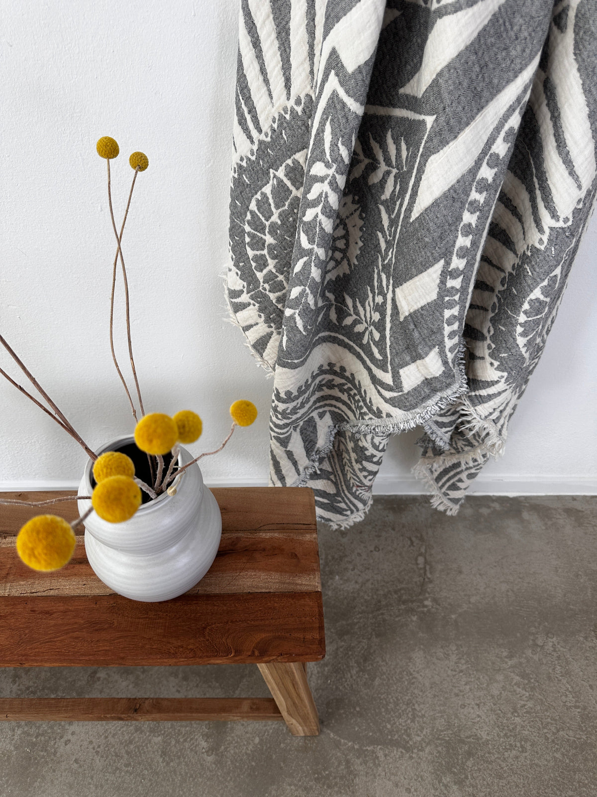 Eine weiße Vase mit gelben Billyball-Blüten steht auf einer Holzbank neben der LEY! CALMA Musselin Decke in Dunkelgrau (2,20 x 2,30 m), deren Boho-Design das graue und weiße Textil auf dem Betonboden ergänzt.