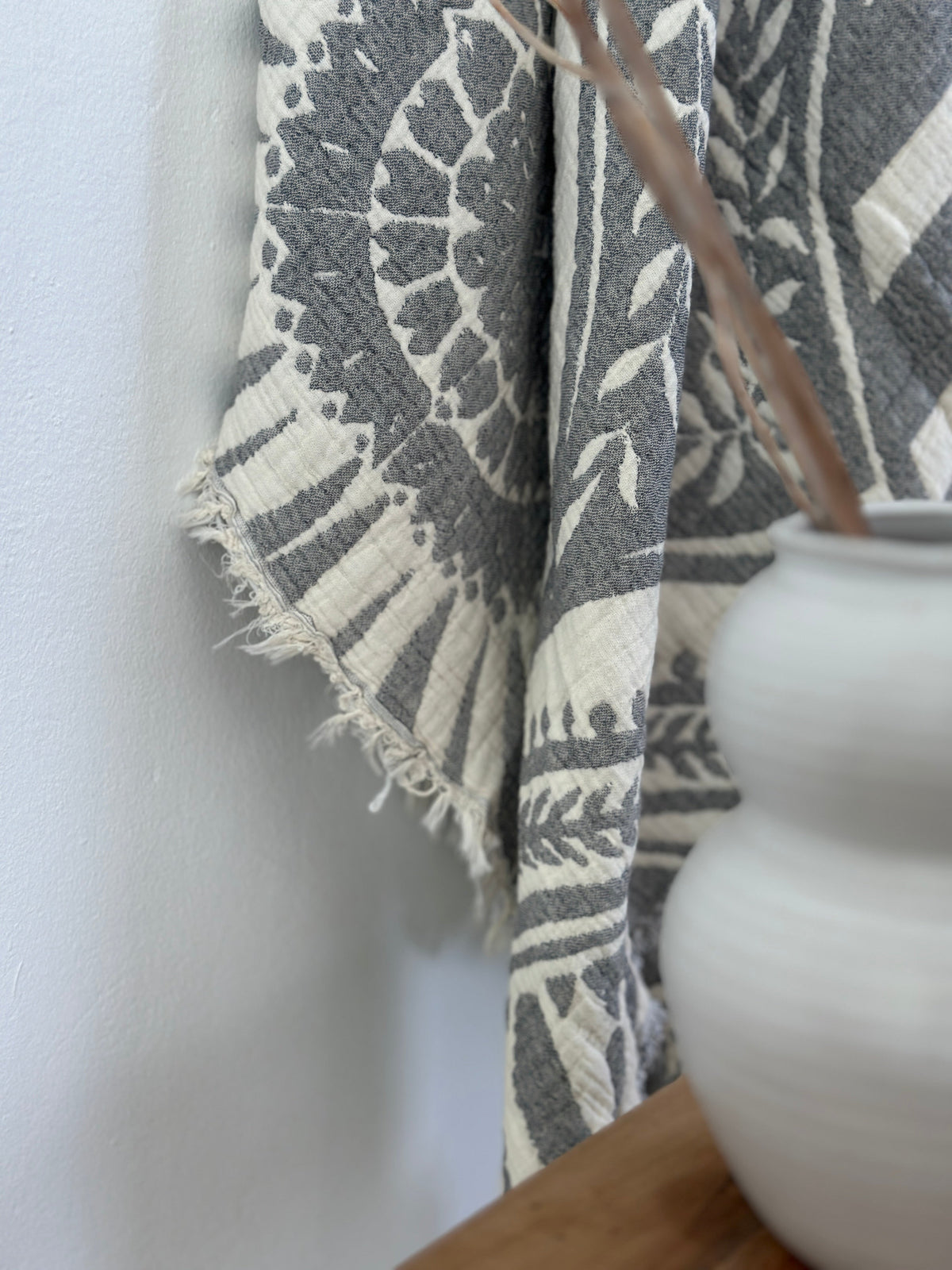 Eine Nahaufnahme der LEY! CALMA Musselin Decke in Dunkelgrau (2,20 x 2,30 m) mit ausgefransten Rändern, drapiert neben einer weißen Keramikvase auf Holz, die ihre weiche Textur und ihr subtiles Boho-Design vor einer hellen Wand hervorhebt.