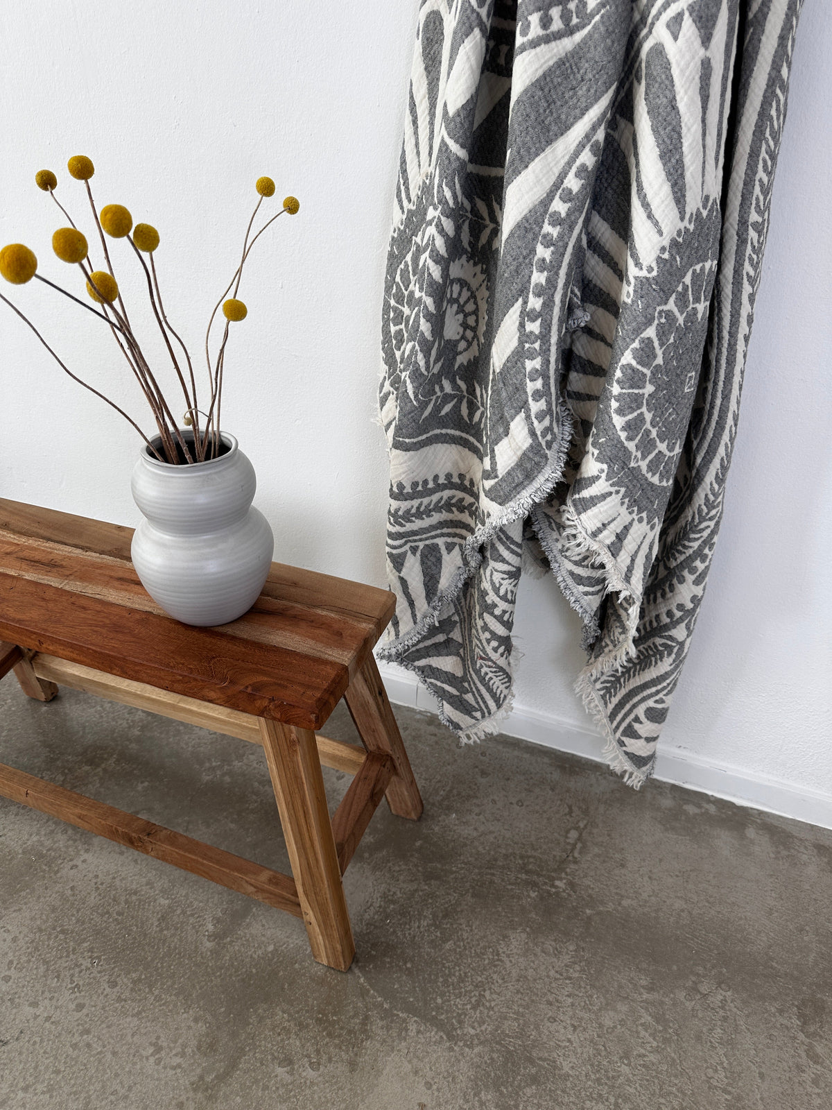 Auf einer Holzbank steht eine graue Vase mit gelben Billyball-Blumen. Daneben hängt die CALMA Musselin Decke in Dunkelgrau von LEY! (2,20 x 2,30 m, Boho Design) hängt an einer weißen Wand über einem Betonboden.