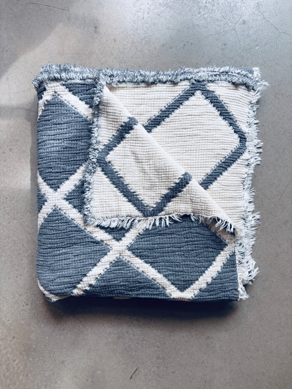 Die gefaltete Musselin Decke von LEY! in Grau-Weiß (2,20 x 2,50 m) mit Fransen liegt auf hellem Beton und verleiht dem Raum einen Hauch Boho-Design.