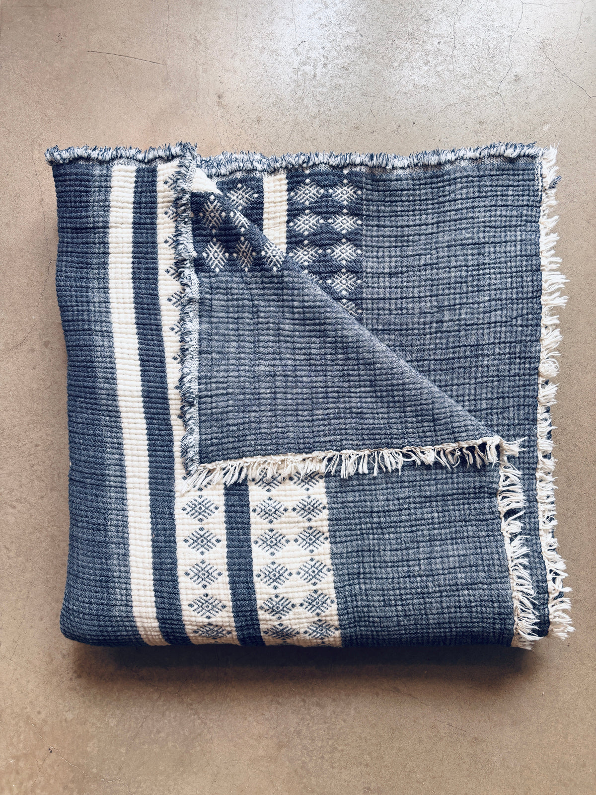 Die LEY! Musselin Decke - Blau-Weiß (2,20 x 2,50 m) mit Fransenkanten und geometrischem Boho-Muster ist auf einer hellbraunen Oberfläche gefaltet, wobei eine Ecke umgeschlagen wird, um die weiche Rückseite freizulegen.