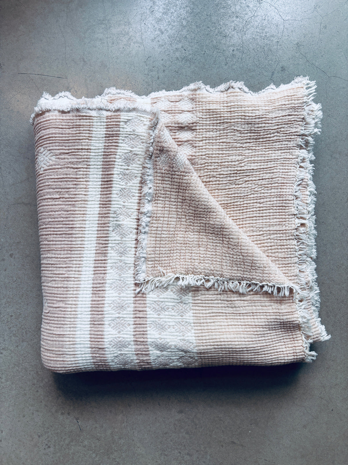 Die gefaltete Musselin Decke - Beige-Weiß von LEY! (2,20 x 2,50 m) im Boho-Design mit Fransen liegt auf einer glatten, hellgrauen Oberfläche.