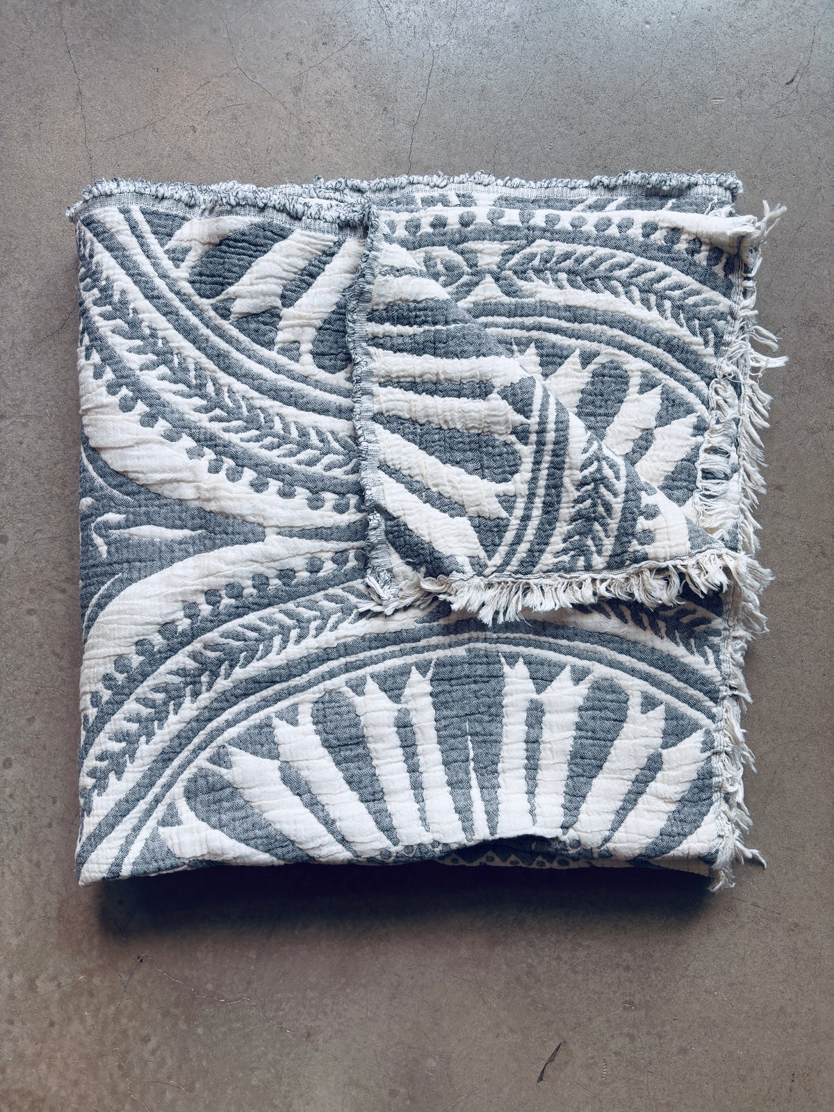 Die CALMA Musselin Decke von LEY! in dunkelgrau (2,20 x 2,30 m) mit stilvollem Boho-Design und ausgefransten Kanten ist auf einer glatten, hellgrauen Oberfläche sauber gefaltet.