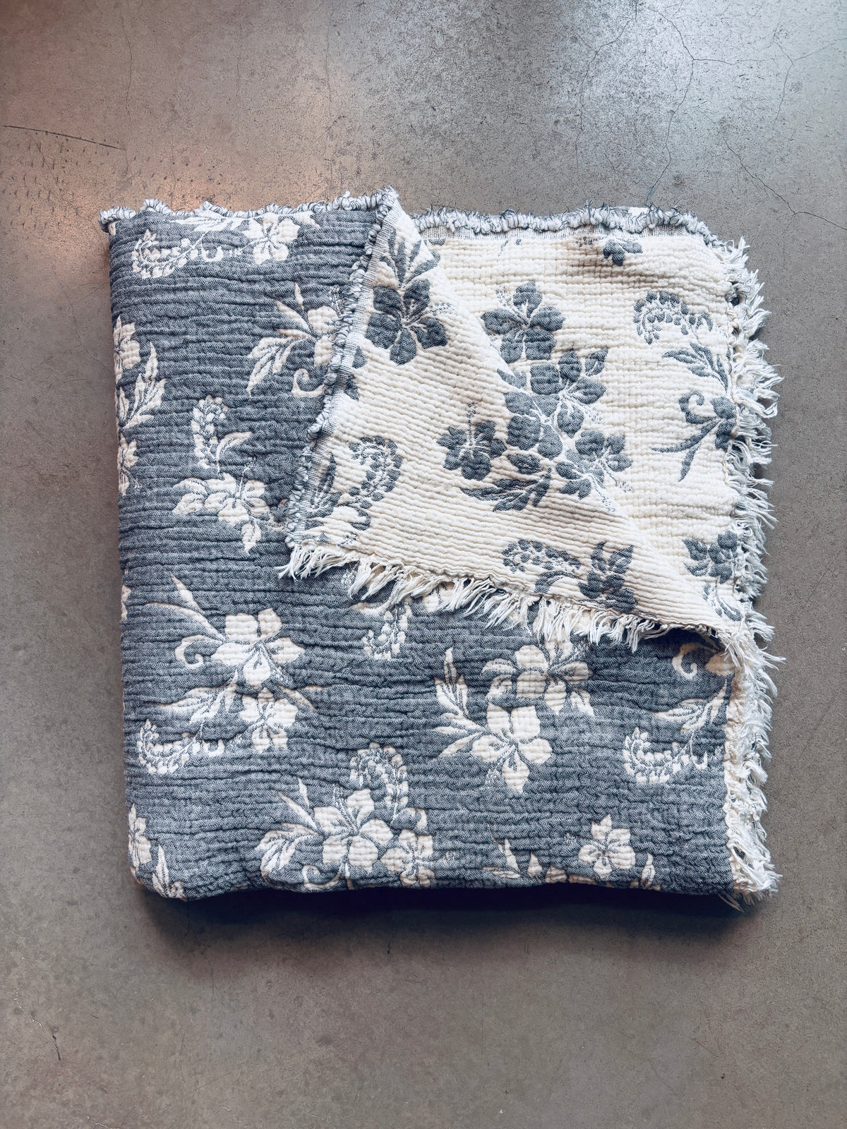 Die LEY! Musselin Decke - ROSE in Grau-Weiß (2,20 x 2,50 m) zeichnet sich durch ein weiches, gefaltetes Design mit weißem, floralem Boho-Muster auf dunklem Stoff aus, das auf der Vorder- und Rückseite auf einer strukturierten grauen Oberfläche abgebildet ist.
