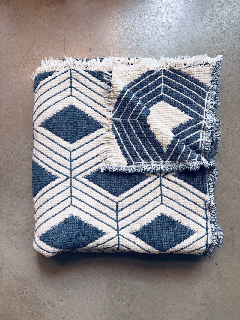 Die LEY! Musselin Decke - Blau-Weiß (2,20 x 2,50 m) mit geometrischem Diamant- und Chevron-Muster sowie Fransen liegt auf einer glatten, hellbraunen Fläche.