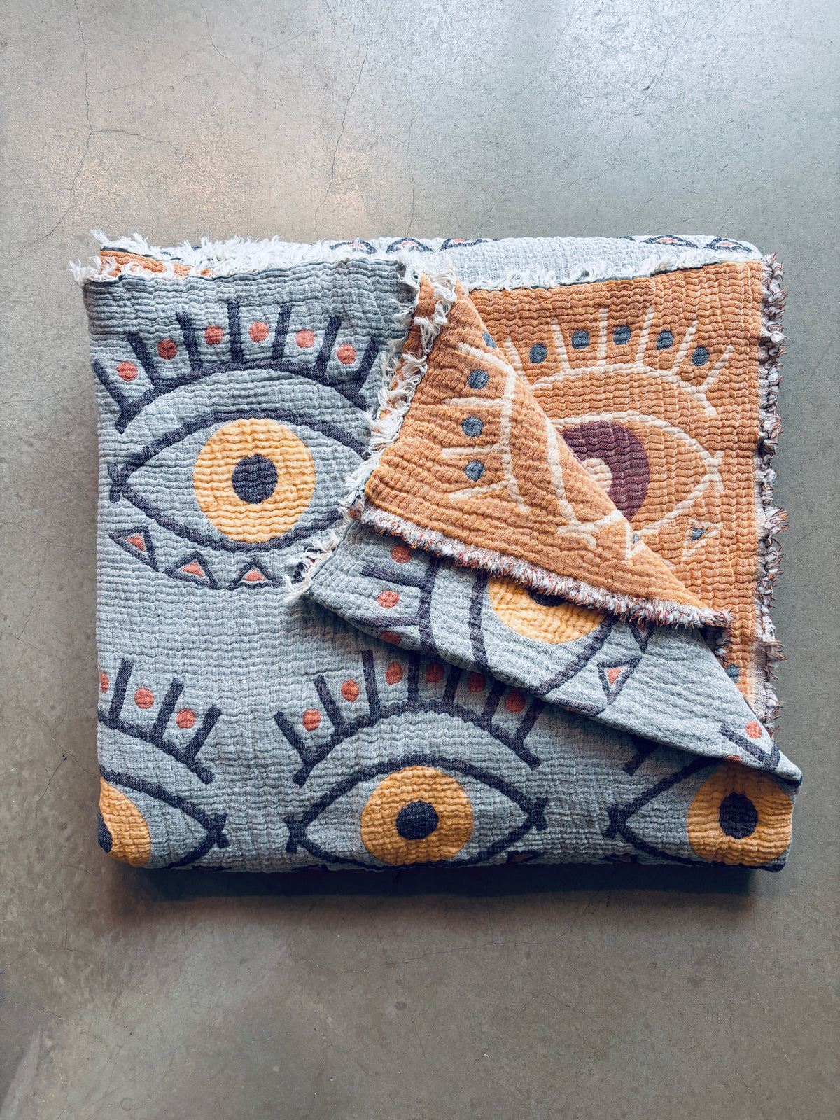 Die Musselin Decke - Vision - Gelb-Blau (2,20 x 2,50 m) von LEY! im Boho-Design mit blauen, gelben und orangenen Augenmustern liegt gefaltet auf einer glatten Fläche; ihr umgeschlagener Rand gibt die orange-lila Rückseite frei - ideal als weiche Decke.