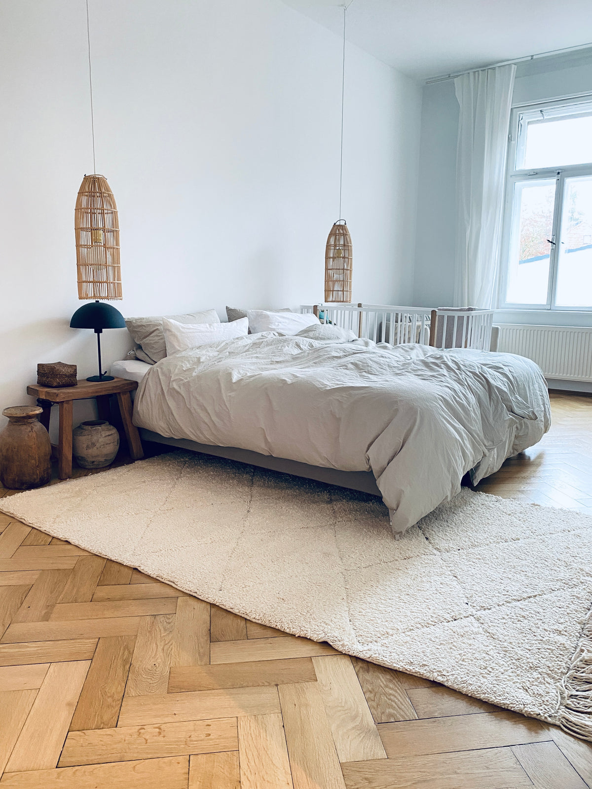 schlafzimmereinrichtung in einer Altbauwohnung. Großes Boxspringbett mit dezenter Bettwäsche. Vor dem Bett ein weißer Berberteppich, ein vintage Hocker als Nachttisch, Rattanlampen am Bett. 