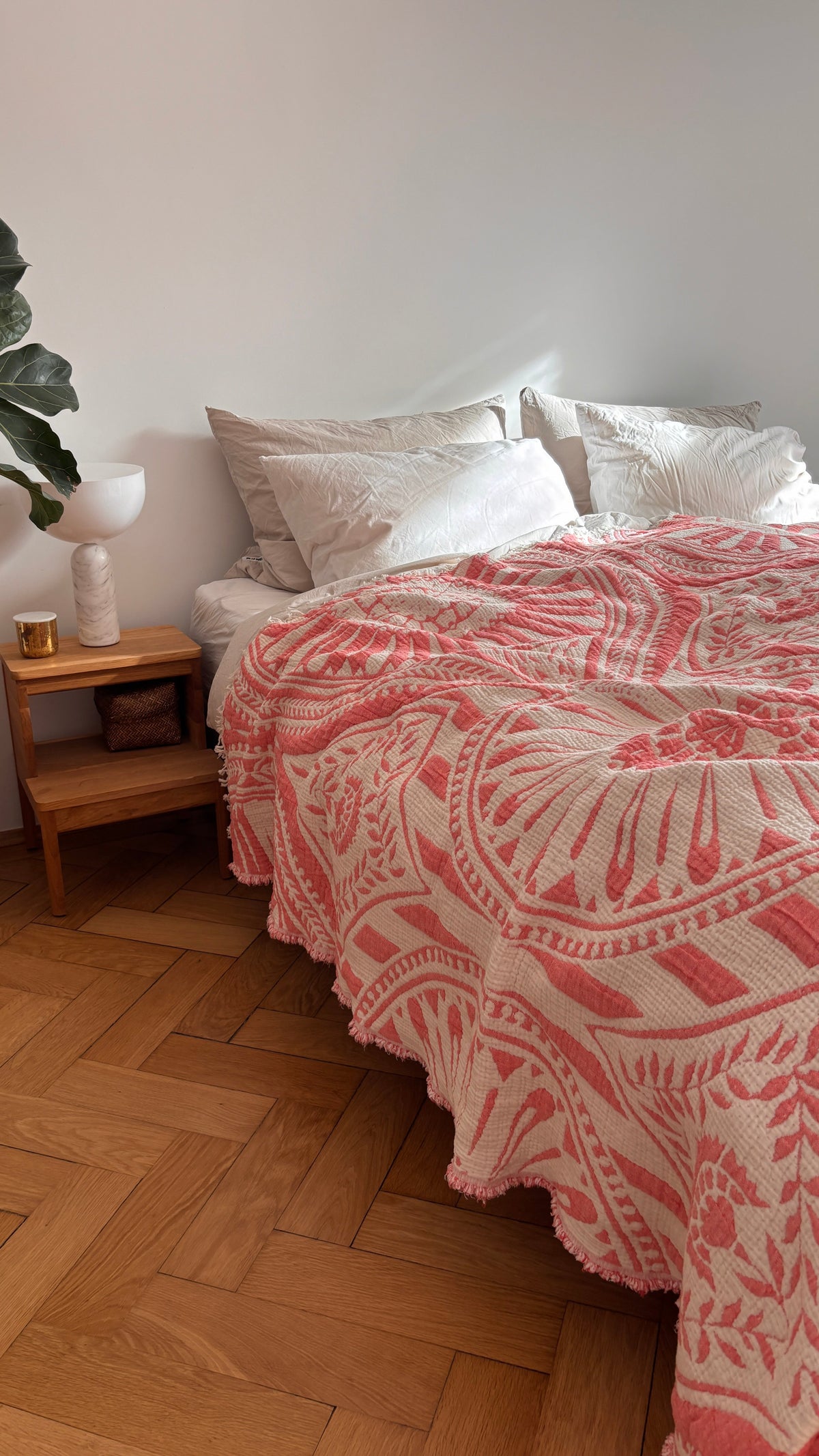 Ein hübsch gemachtes Bett mit weißen Kissen und der CALMA Musselin Decke - Koralle (2,20 x 2,30 m) von LEY! sorgt für Boho-Stimmung neben einem hölzernen Nachttisch mit Kerze, Korb und Lampe. Das Zimmer hat einen Holzboden, weiße Wände und eine Blattpflanze in der Ecke.