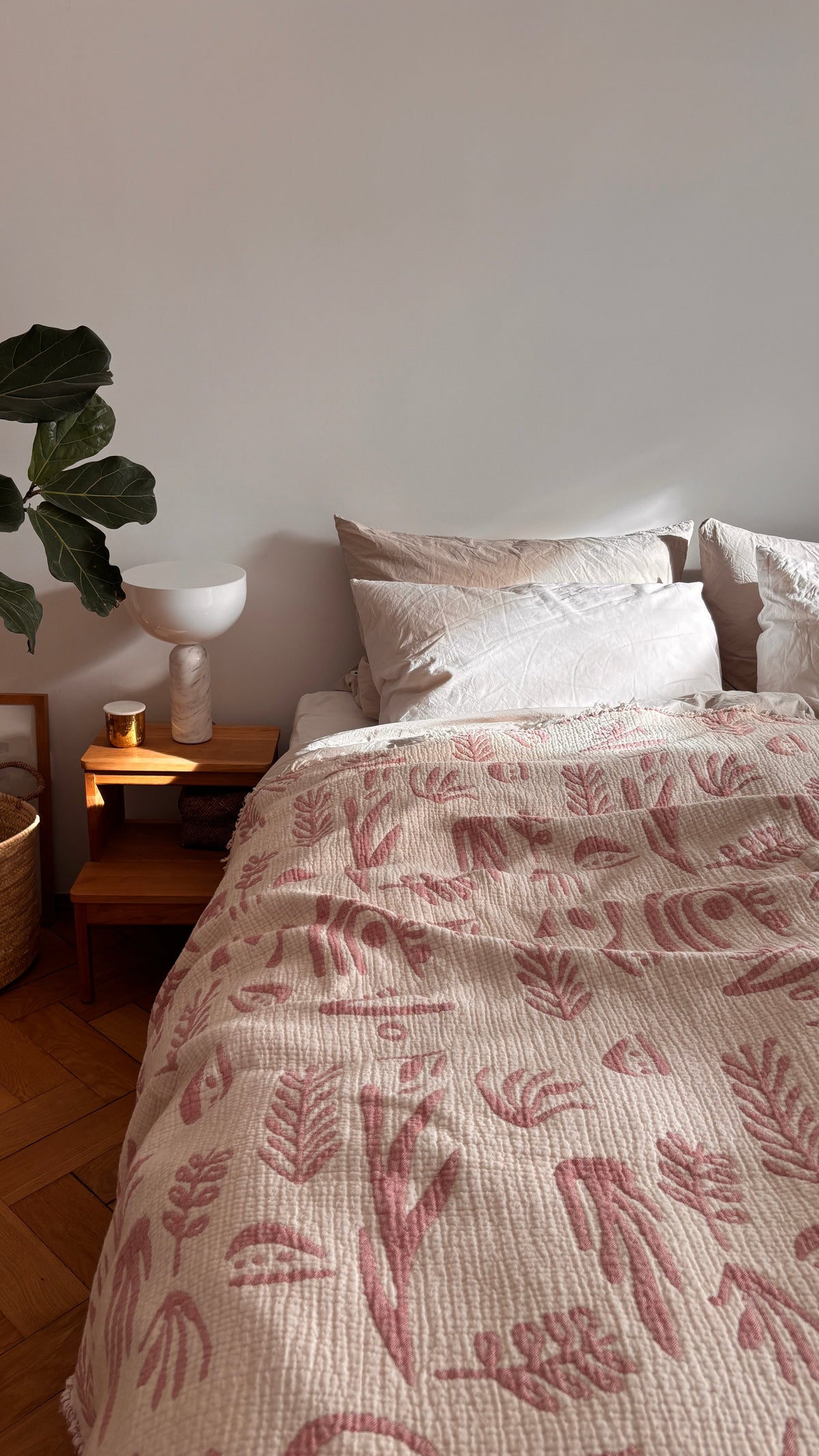 Ein gemütliches Bett mit weißen Kissen und der LEY! CALMA Musselin Decke in Rosé-Weiß (2,30 x 2,40 m) steht neben einem hölzernen Nachttisch mit einer Pflanze, einer Lampe, einer Kerze und einem Foto in einem luftigen, sanft beleuchteten, minimalistischen Schlafzimmer mit Boho-Akzenten.