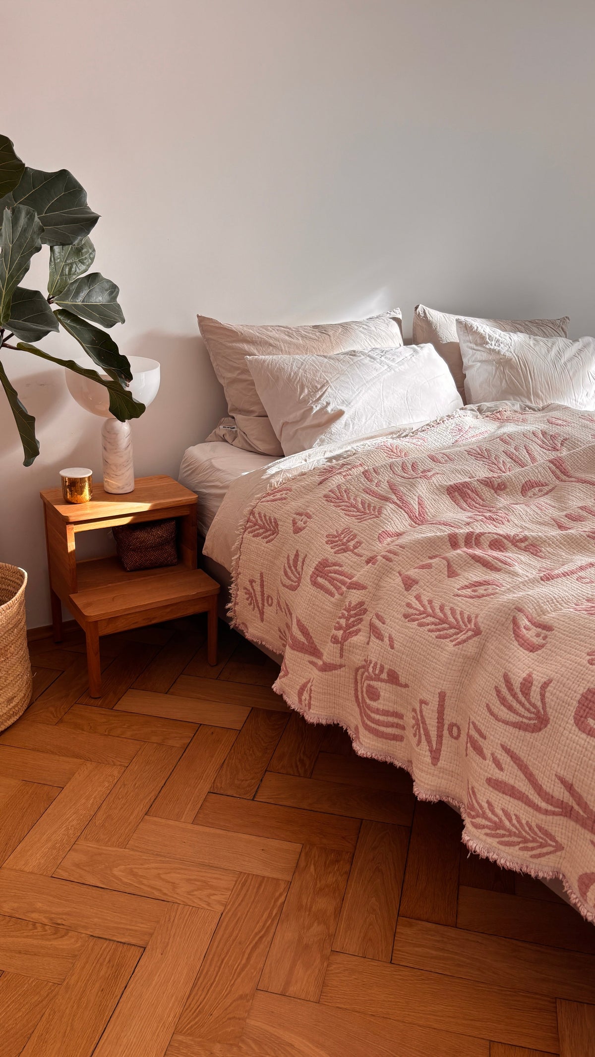Ein gemütliches Schlafzimmer bietet die CALMA Musselin Decke - Rosé-Weiß (2,30 x 2,40 m) von LEY! mit atmungsaktivem Komfort und Boho-Design. Weiße Kissen, ein Holznachttisch mit Lampe und Kerze, eine Topfpflanze und ein geflochtener Korb machen den Look komplett.