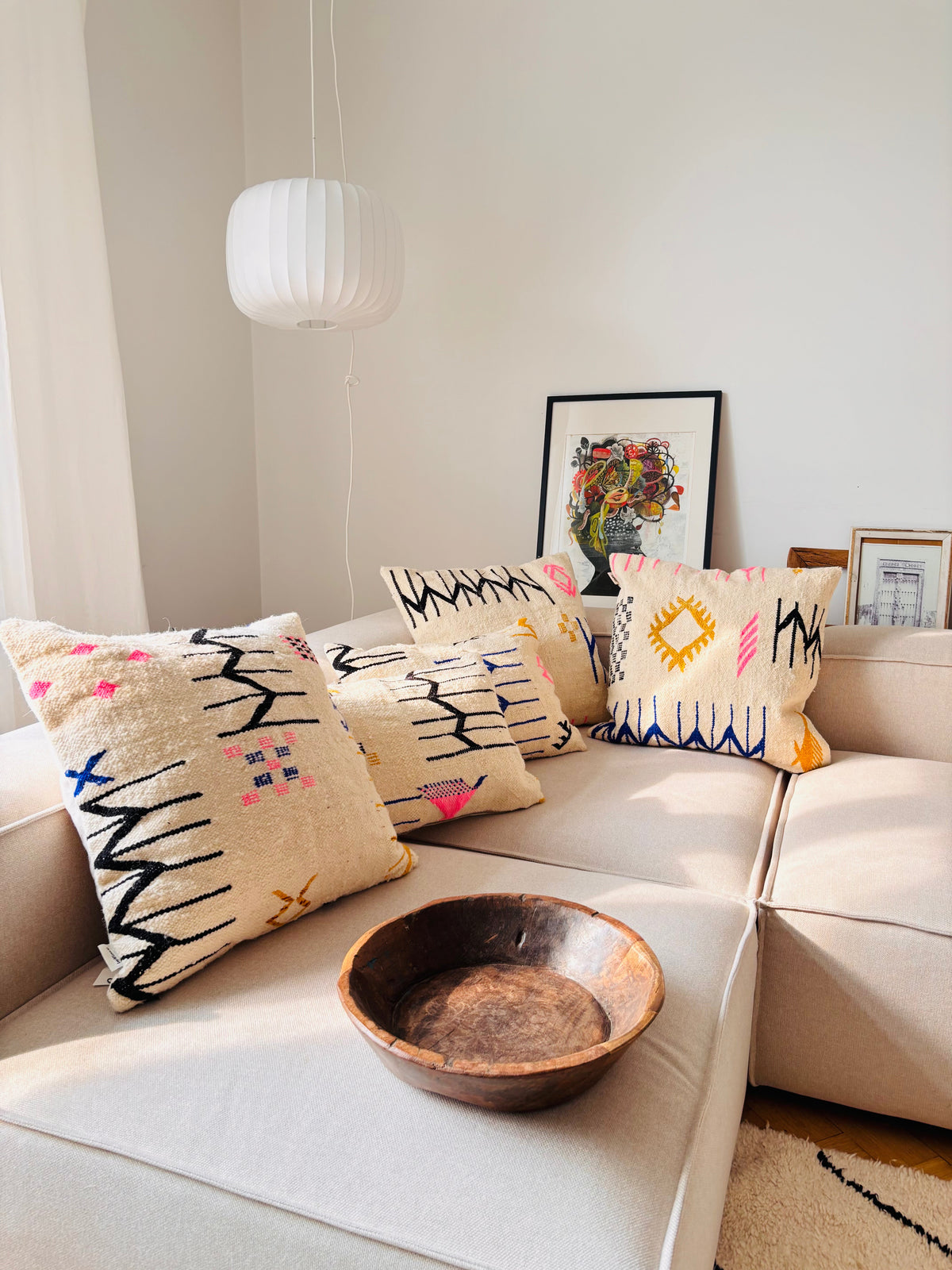 Beigefarbenes Sofa mit einem LEY! Kissen - Colorful Line - 60x60, Holzschale, gerahmter Kunst und weißer Pendelleuchte im Sonnenlicht.